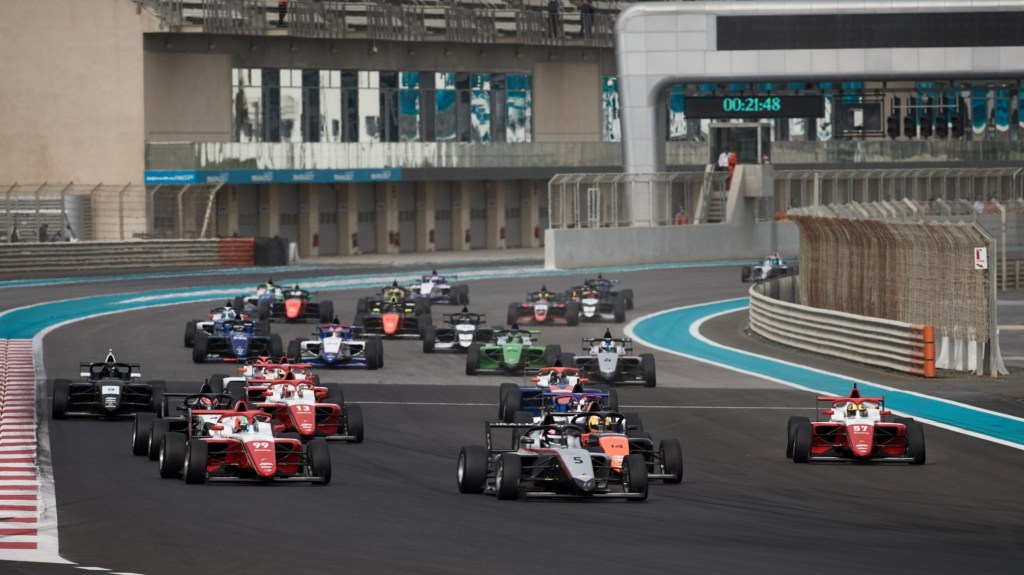 A group of open-wheel cars prepares to turn left from the main straight at Yas Marina Circuit, where 21:48 is visible in the background on the timing clock