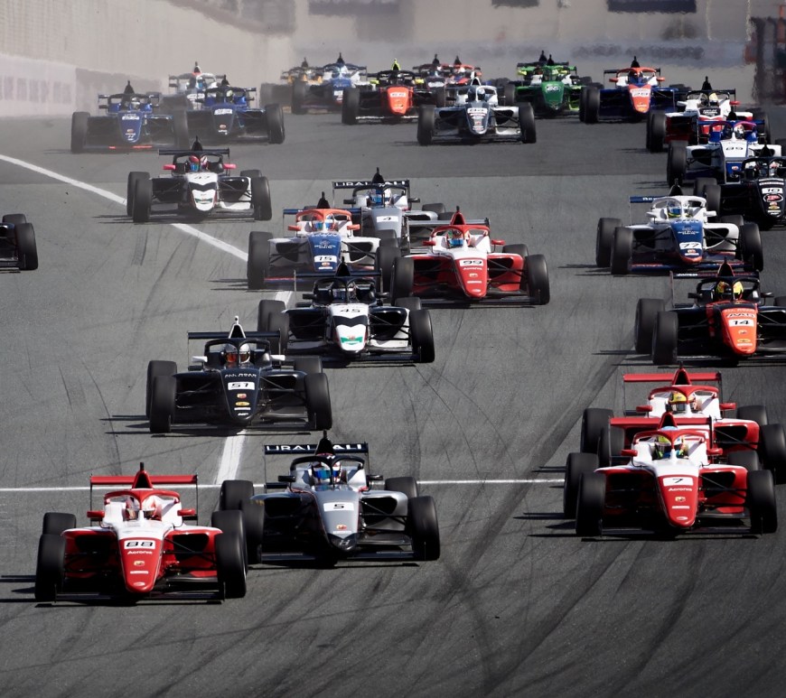 A group of 25+ open-wheel cars heads downhill on a straight