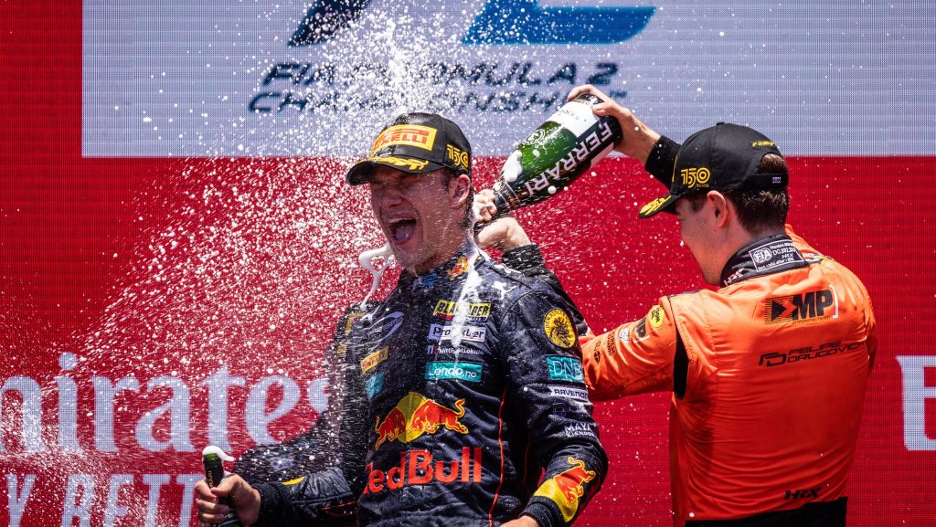 Hauger celebrating on the podium with man wearing orange spraying champagne down his back