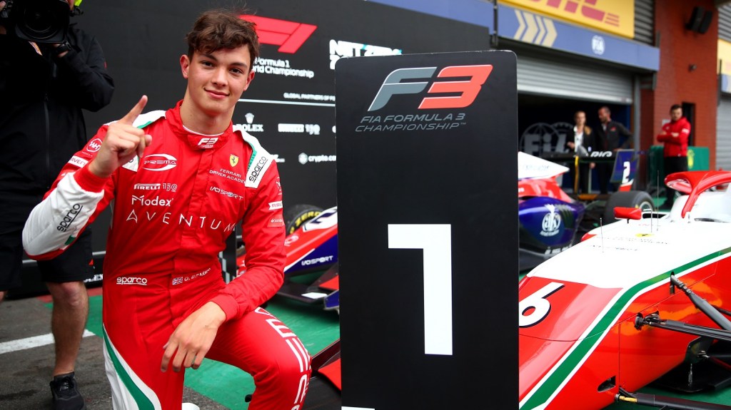 Man in red PREMA racing suit standing beside a #1 sign and his car with his finger raised to represent first place.