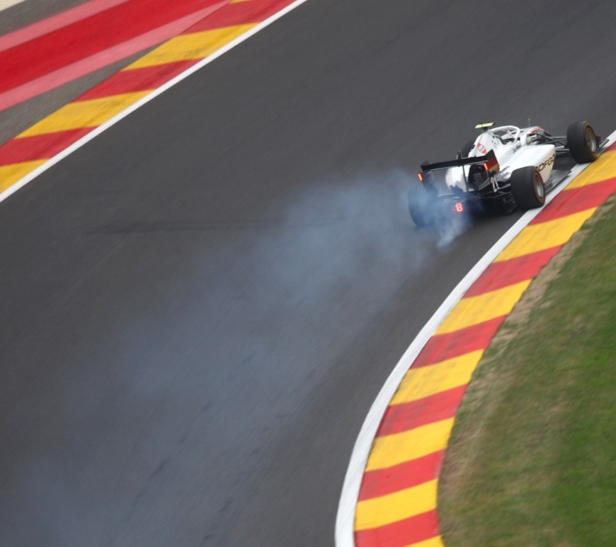 White formula style F3 car travelling uphill and to the right at high speed with smoke emanating from underneath the car.