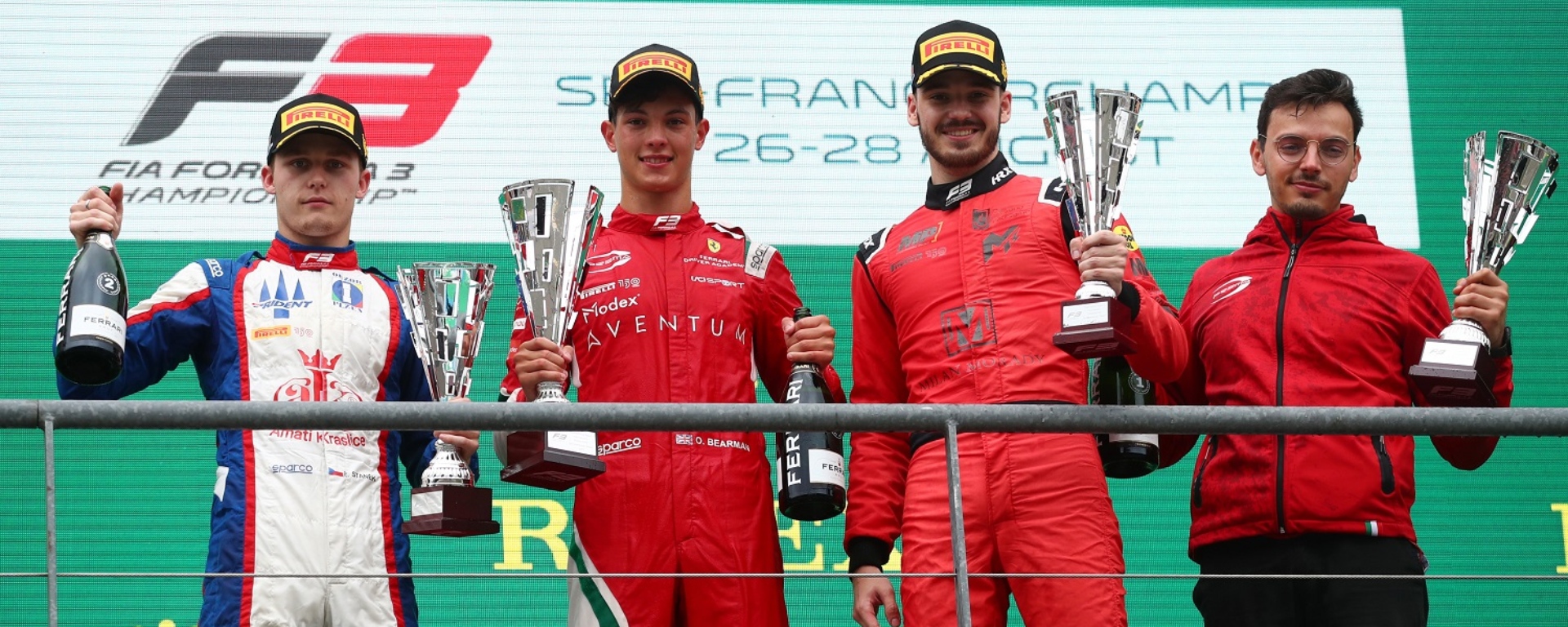 4 people on the podium, Roman Stanek (far left) in white suit, Ollie Bearman (middle left) in red suit, Alexander Smolyar (middle right) in red suit, and a Prema team member (far right) in a red jumper, all holding trophies.