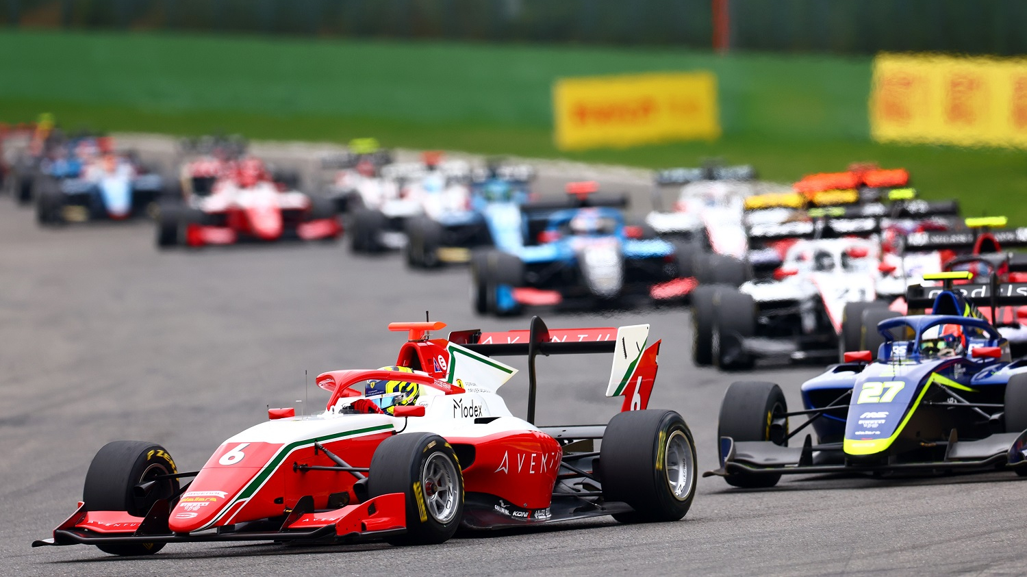 Red and white formula style car driving to the left of the camera with many cars behind it.