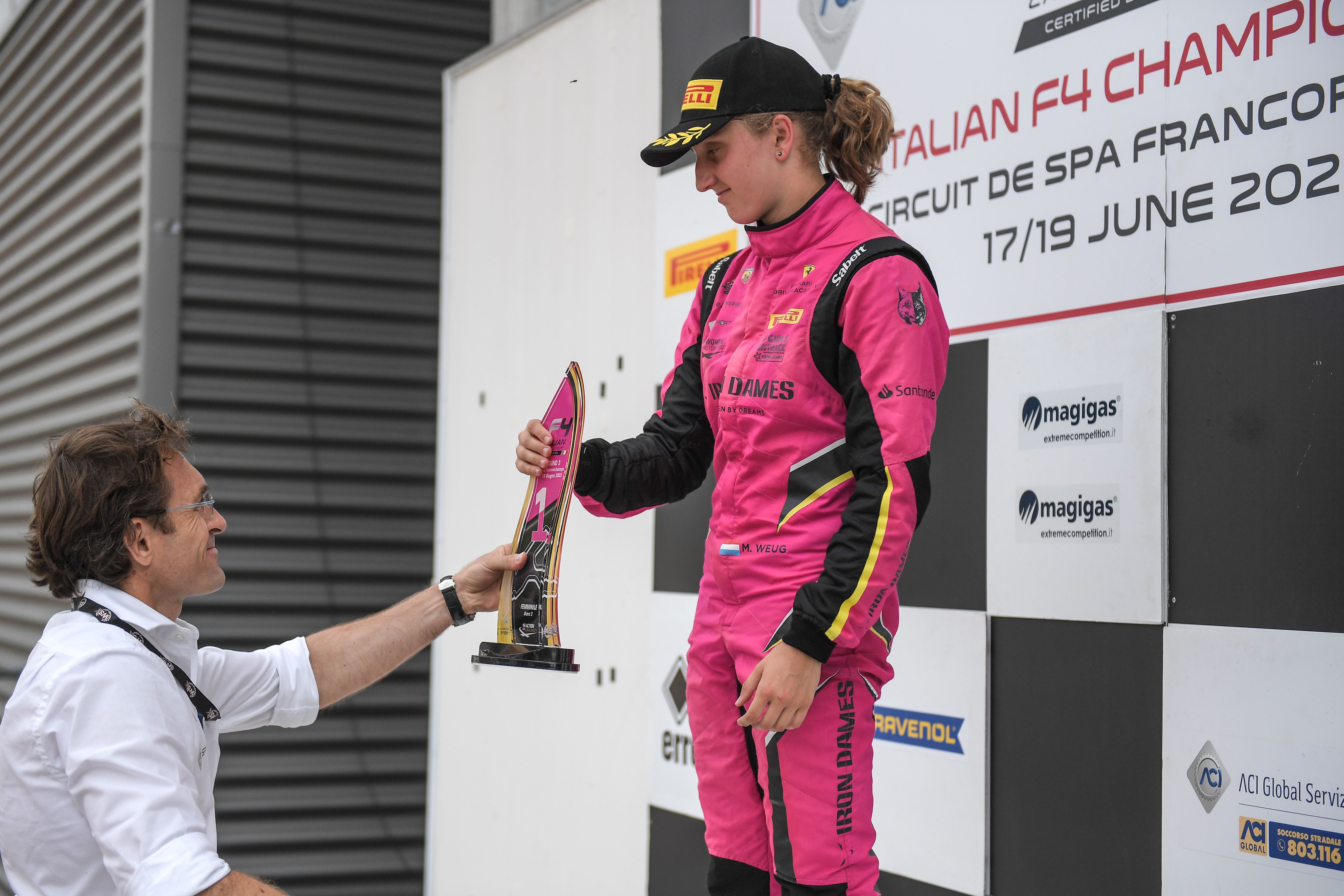 A man in a white button-down shirt hands a transparent gold trophy to Maya Weug in pink and black racing overalls and a black cap with the Pirelli logo. The trophy is emblazoned with a #1; the Spa-Francorchamps circuit layout; the FIA Action for Road Safety, ACI Sport and WSK promotions logos;and the inscriptions 'Round 3, Spa-Francorchamps, 19 Giugno 2022' and 'Femminile, Gara 2'.