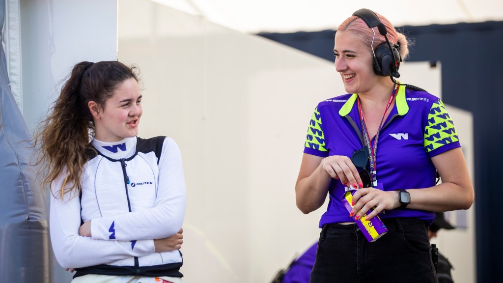 A young woman with brown hair and a white racing vest with purple 'W' insignias chats to another woman with pink dyed hair, a black headset, a purple and neon green W Series top and flourescent pink nail polish with the outline of palm trees in black