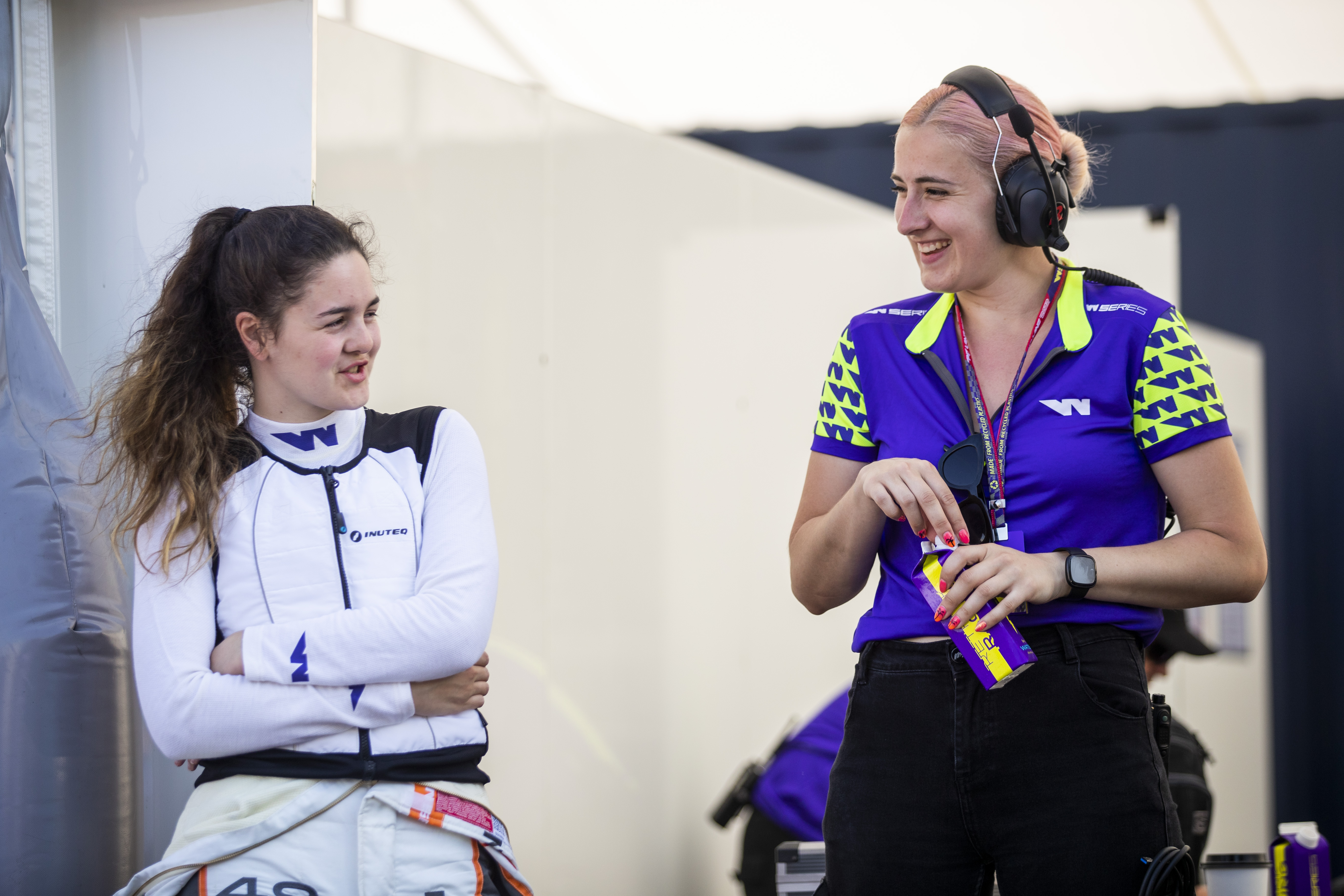 A young woman with brown hair and a white racing vest with purple 'W' insignias chats to another woman with pink dyed hair, a black headset, a purple and neon green W Series top and flourescent pink nail polish with the outline of palm trees in black