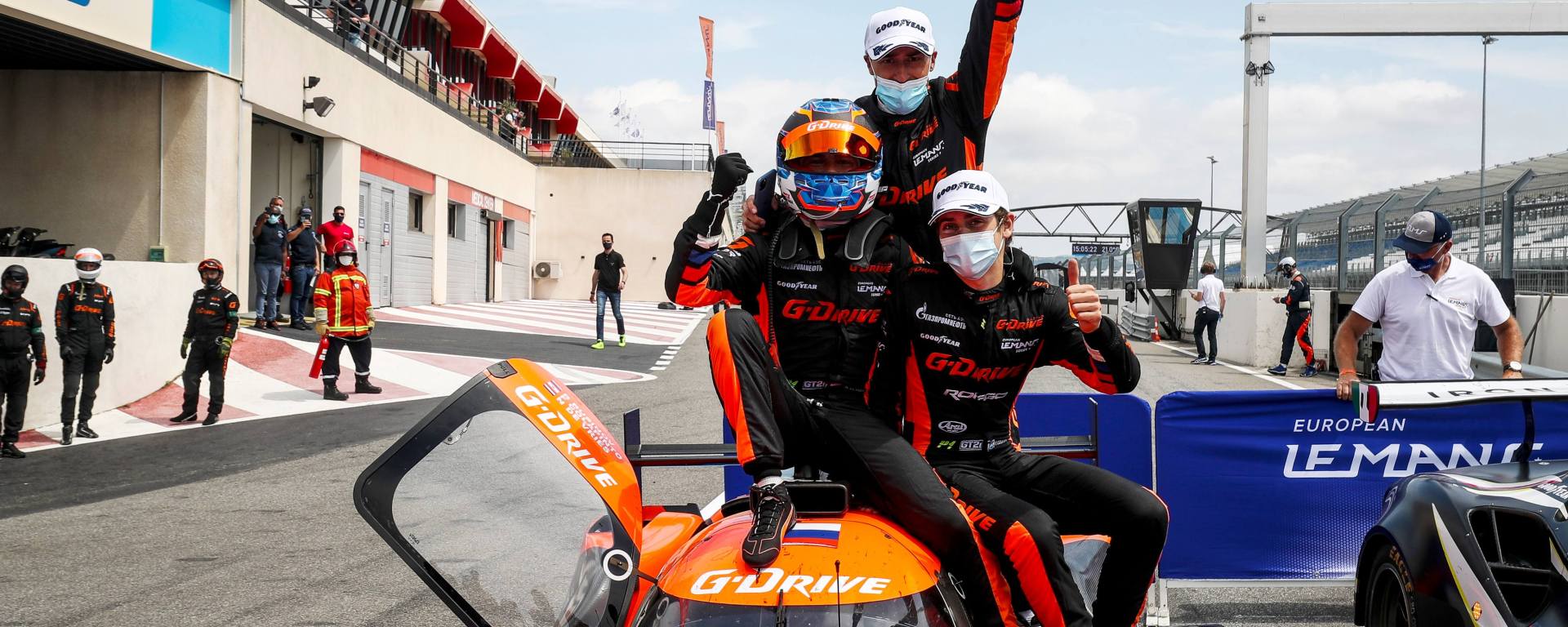 3 male drivers in black and orange suits sitting on top of orange prototype car celebrating with thumbs up and arms in the air.