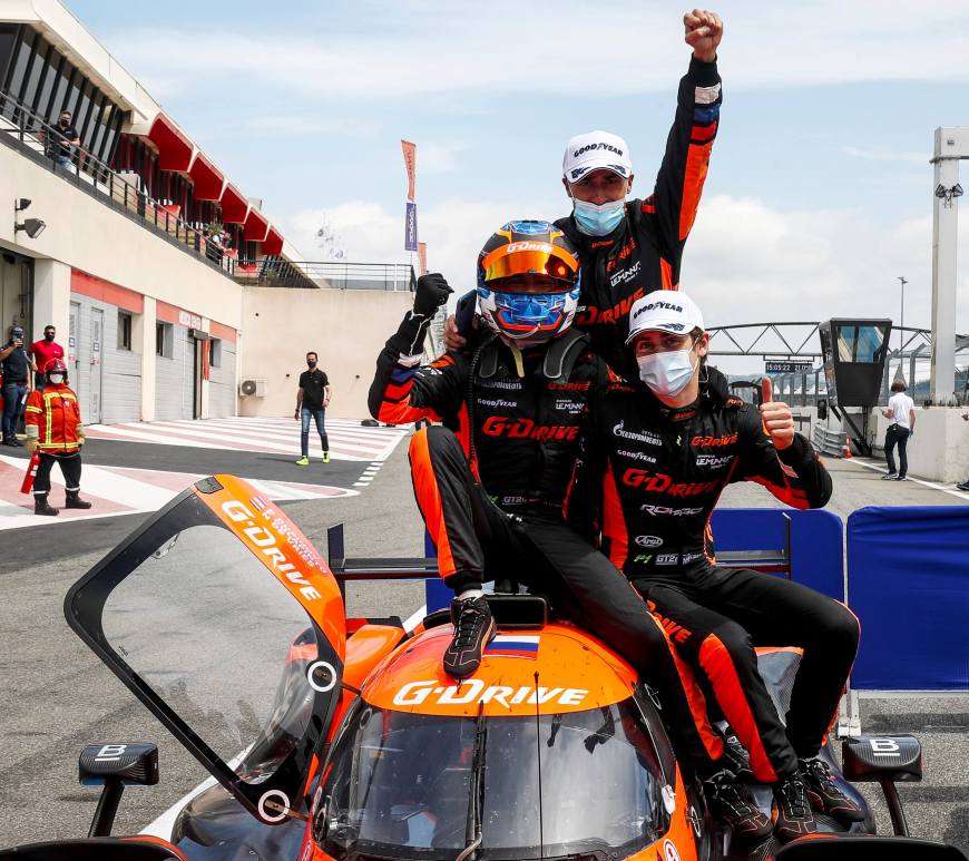 3 male drivers in black and orange suits sitting on top of orange prototype car celebrating with thumbs up and arms in the air.