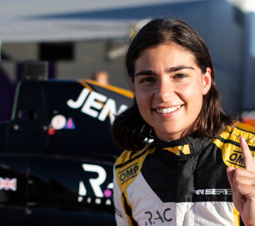 A woman in a black, white and gold racing suit squats in front of her racecar and holds her left index finger in the air