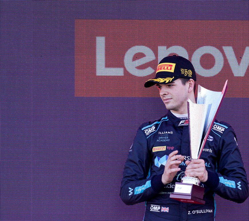 A young man (Zak O'Sullivan) in a black Pirelli 150th anniversary cap and a navy and teal racing suit holds the silver-hued second-place trophy on the podium. There is a purple screen behind him with the Lenovo logo in red.