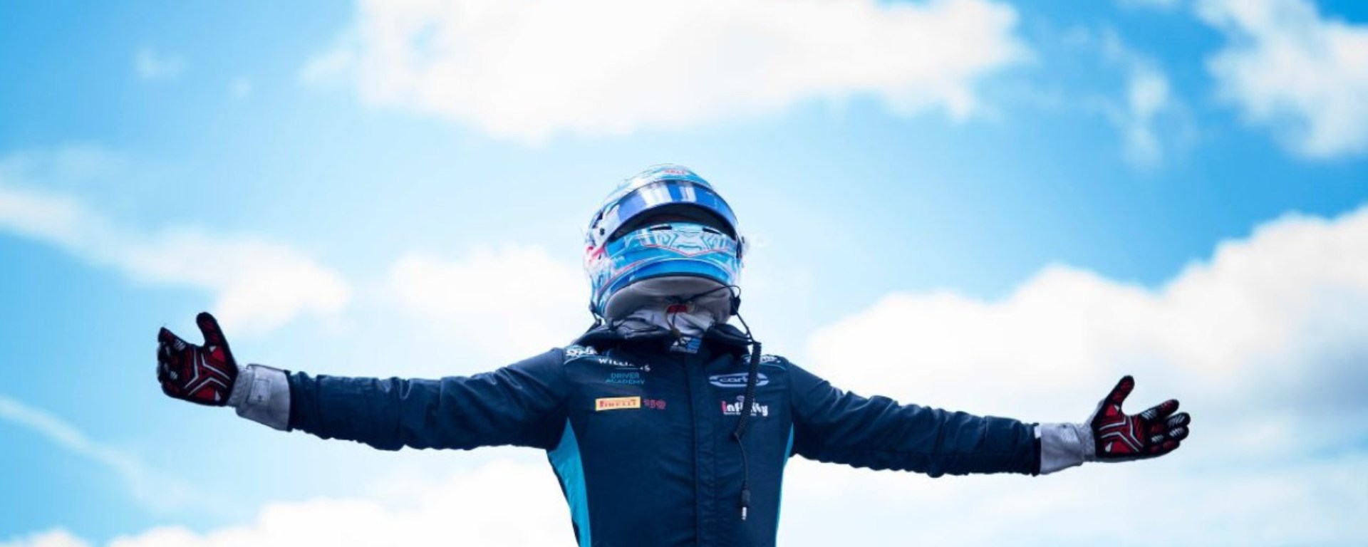 Man (Logan Sargeant) in navy and light blue racing suit and helmet standing with arms spread open with blue sky and clouds behind him.