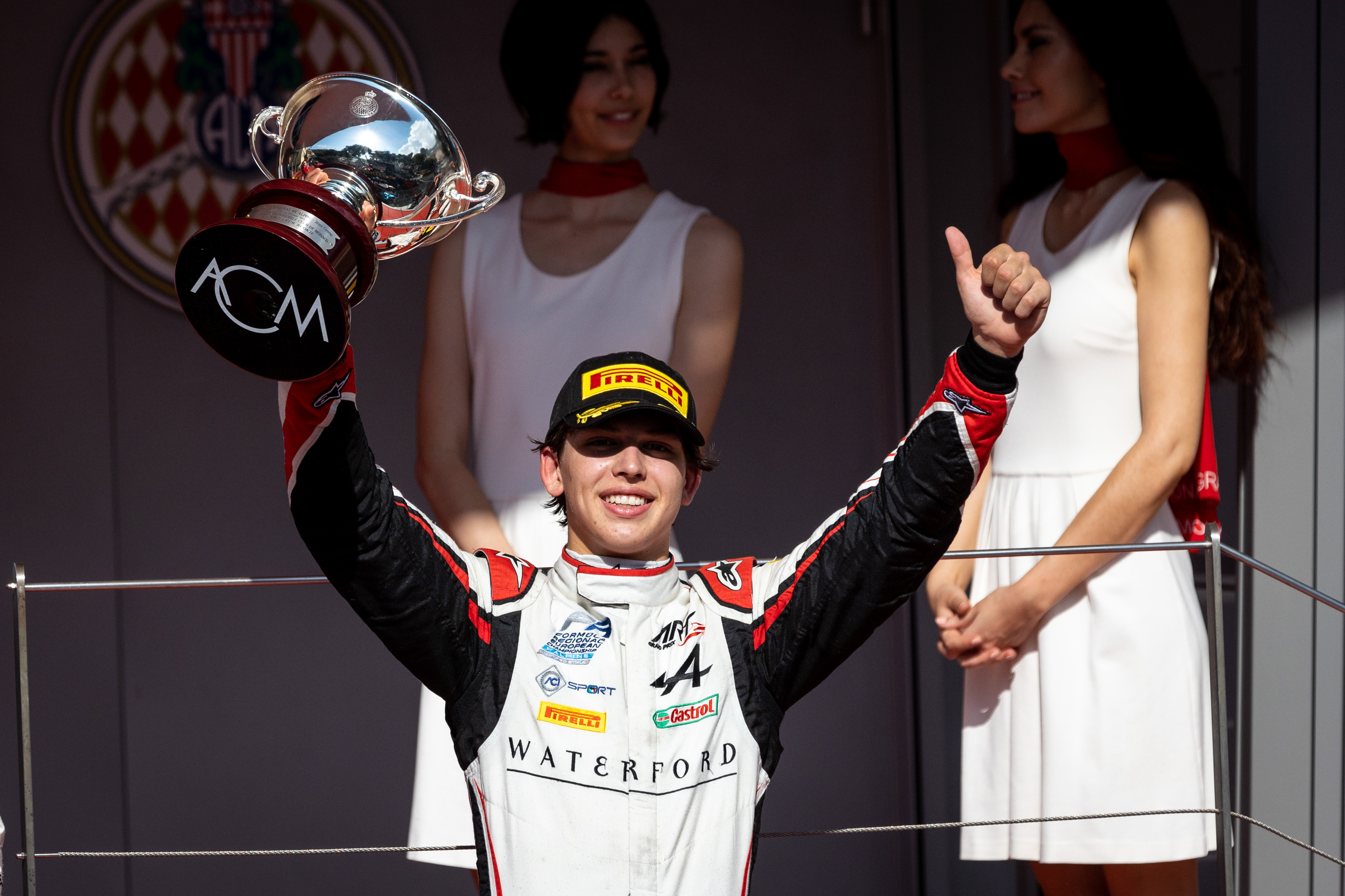 On the podium after FRECA's second race at Monaco, a smiling Laurens van Hoepen gives a thumbs-up gesture with his left hand and holds the trophy for best rookie, engraved with the Automobile Club de Monaco logo on its base, aloft in his right hand.
