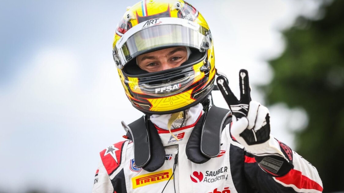 A driver in a yellow, black and red helmet gives a peace sign to the camera with his left hand