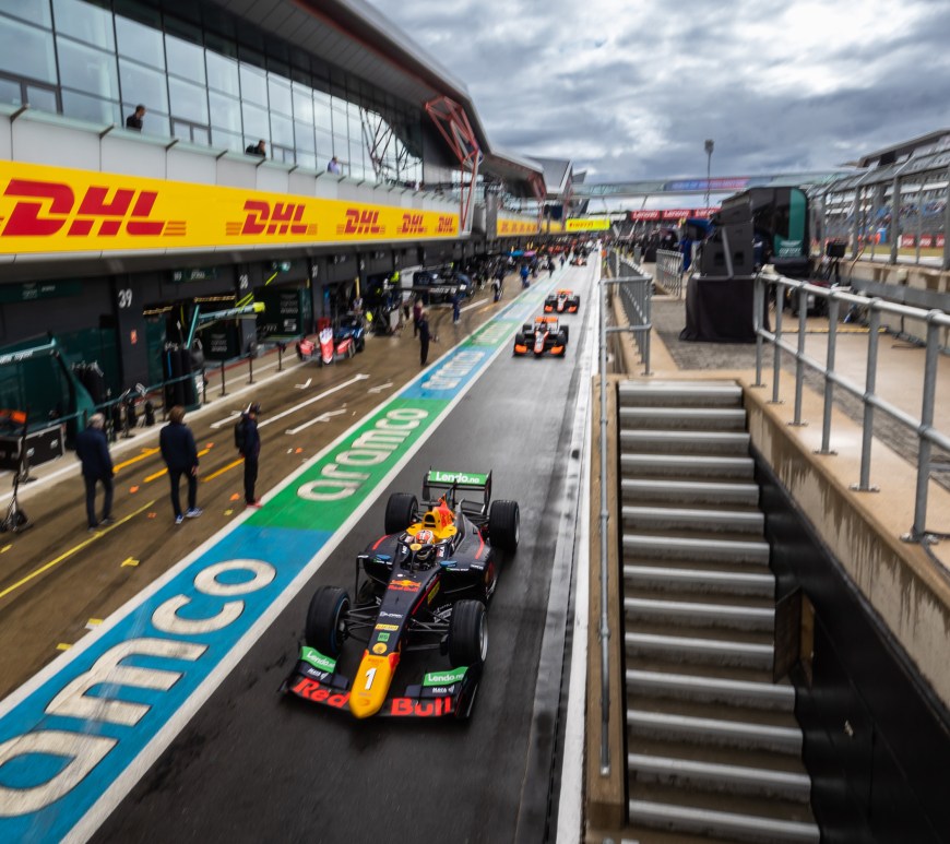 A blue, yellow, red and green racecar drives toward the camera in the pit lane of the Silverstone Circuit. To the left are the team garages and a stripe of green and blue aramco branding; to the right is the stairwell with a trackside viewing area for the pitwall; and behind Hauger are the two black and orange Van Amersfoort Racing cars of Jake Hughes and David Beckmann.