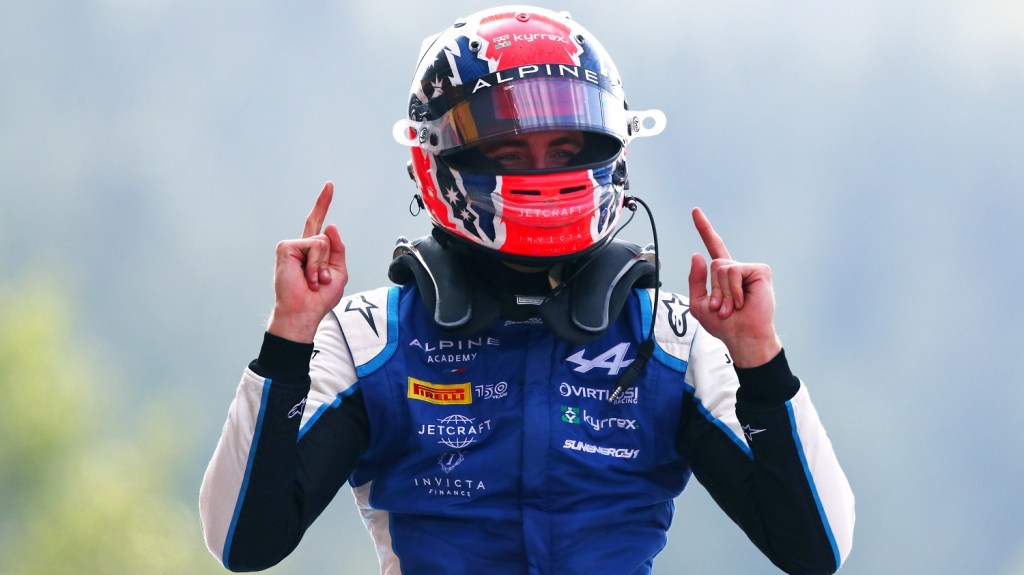 Man in black, white, and blue racing suit with red and blue helmet holds both ands up with index fingers in the air celebrating his first place.