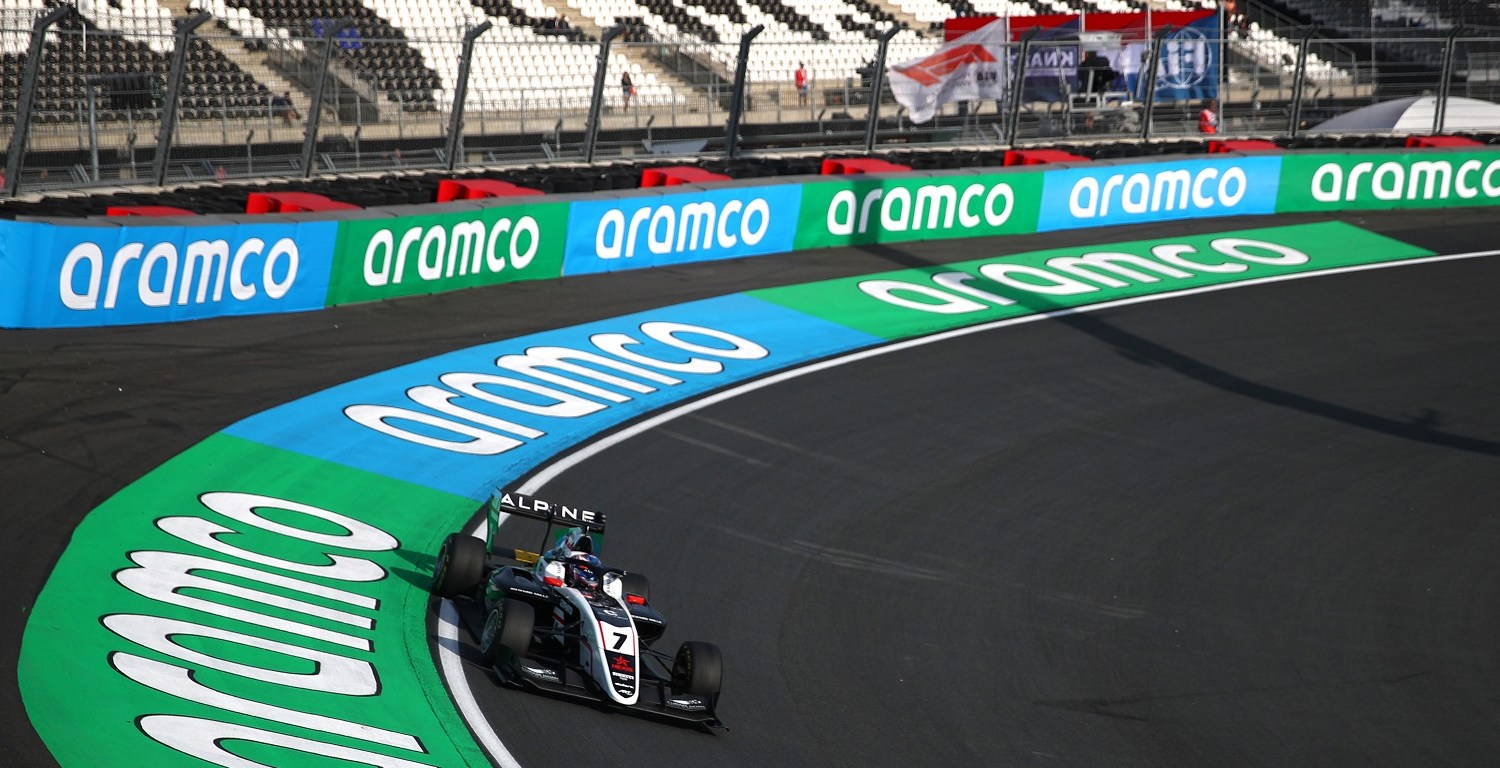 The white and black racecar of Victor Martins takes the high line through Zandvoort's banked left-hander, Turn 3. The right-rear wheel of the car is touching the white line demarcating the edge of the circuit. There is Aramco signage surrounding the track on the wall and on the asphalt.