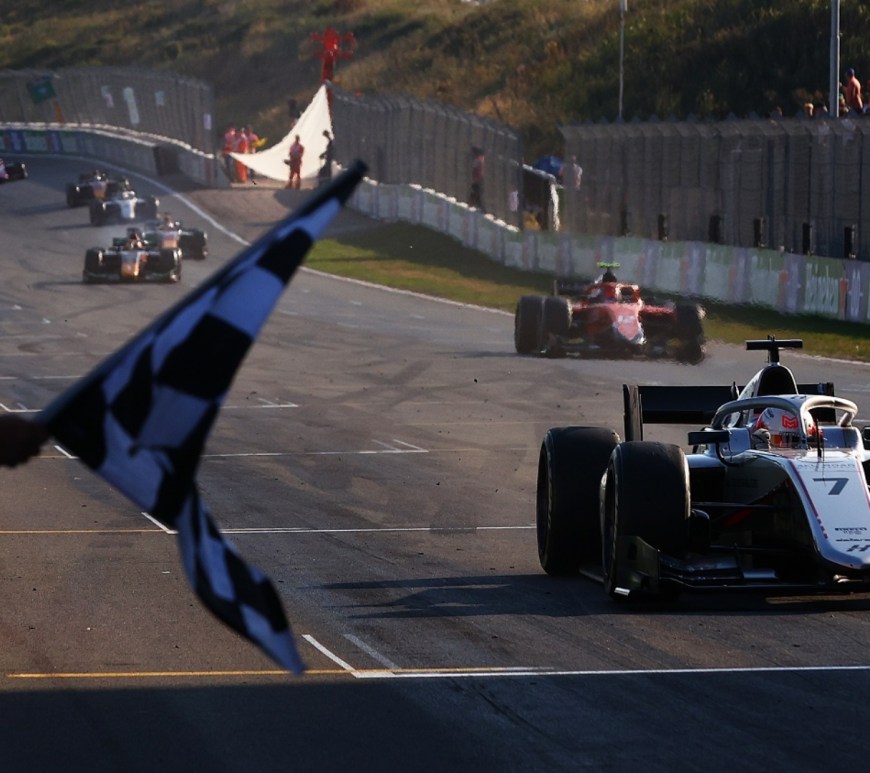 Silver formula 2 car driving towards the camera with the chequered flag waiving and the rest of the cars behind.