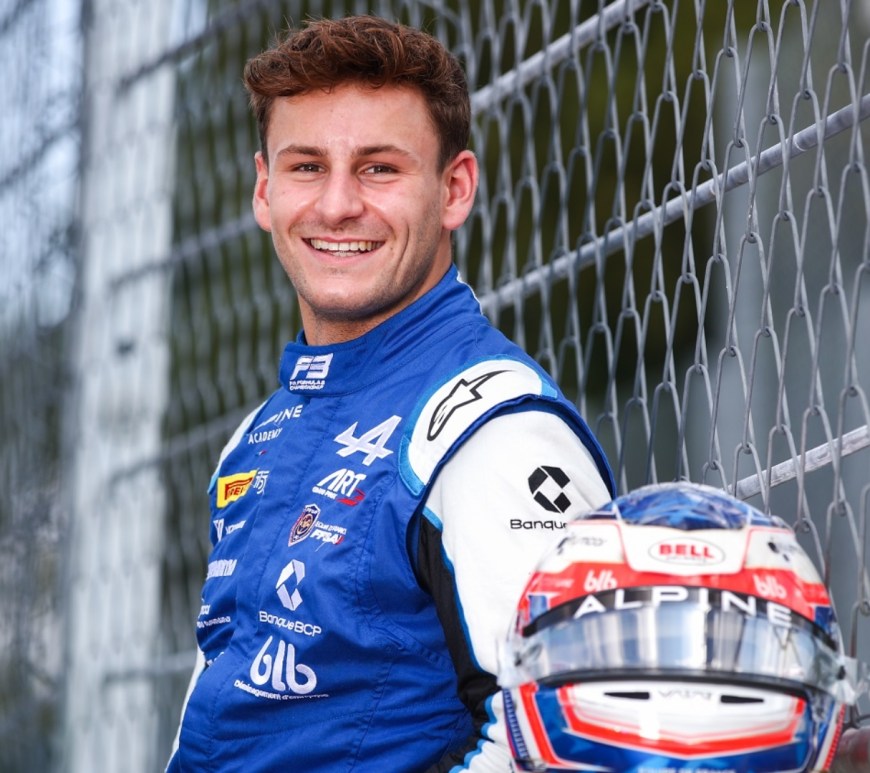 Man in blue and white racing suit standing facing to the left of the camera with head turned smiling at camera.