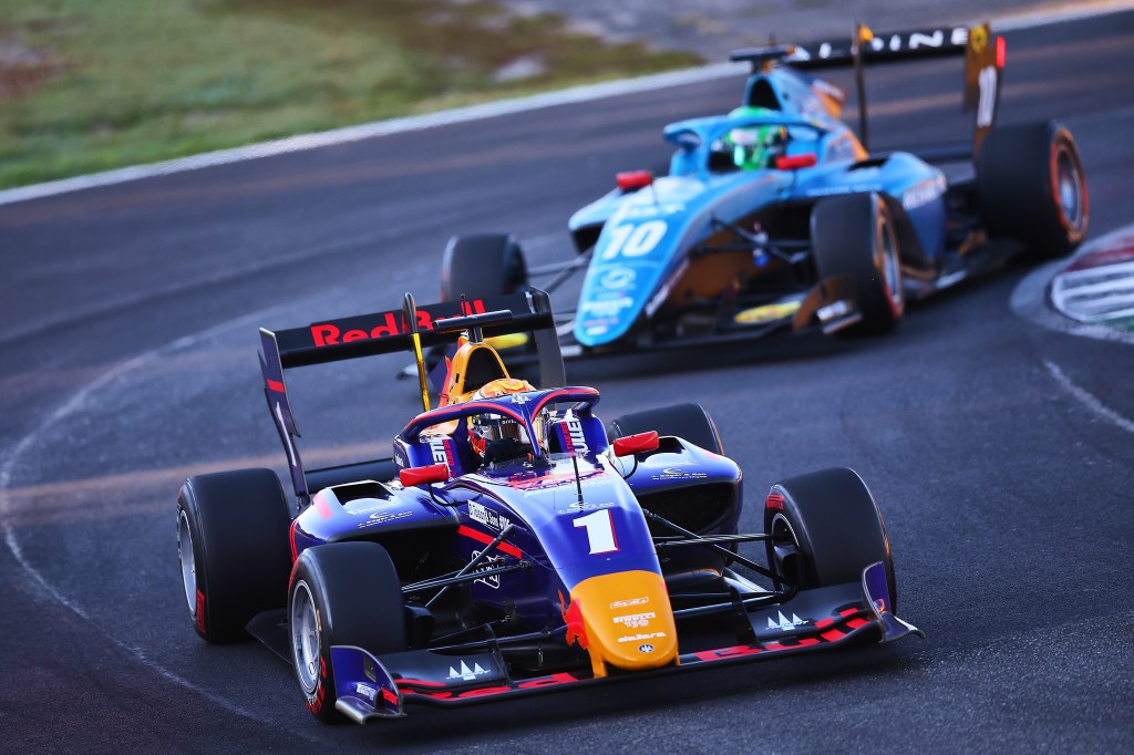 A blue, gold and red racecar with Red Bull branding, driven by Jonny Edgar, makes a left turn. A teal racecar driven by Caio Collet, wearing a bright green crash helmet, pursues Edgar.