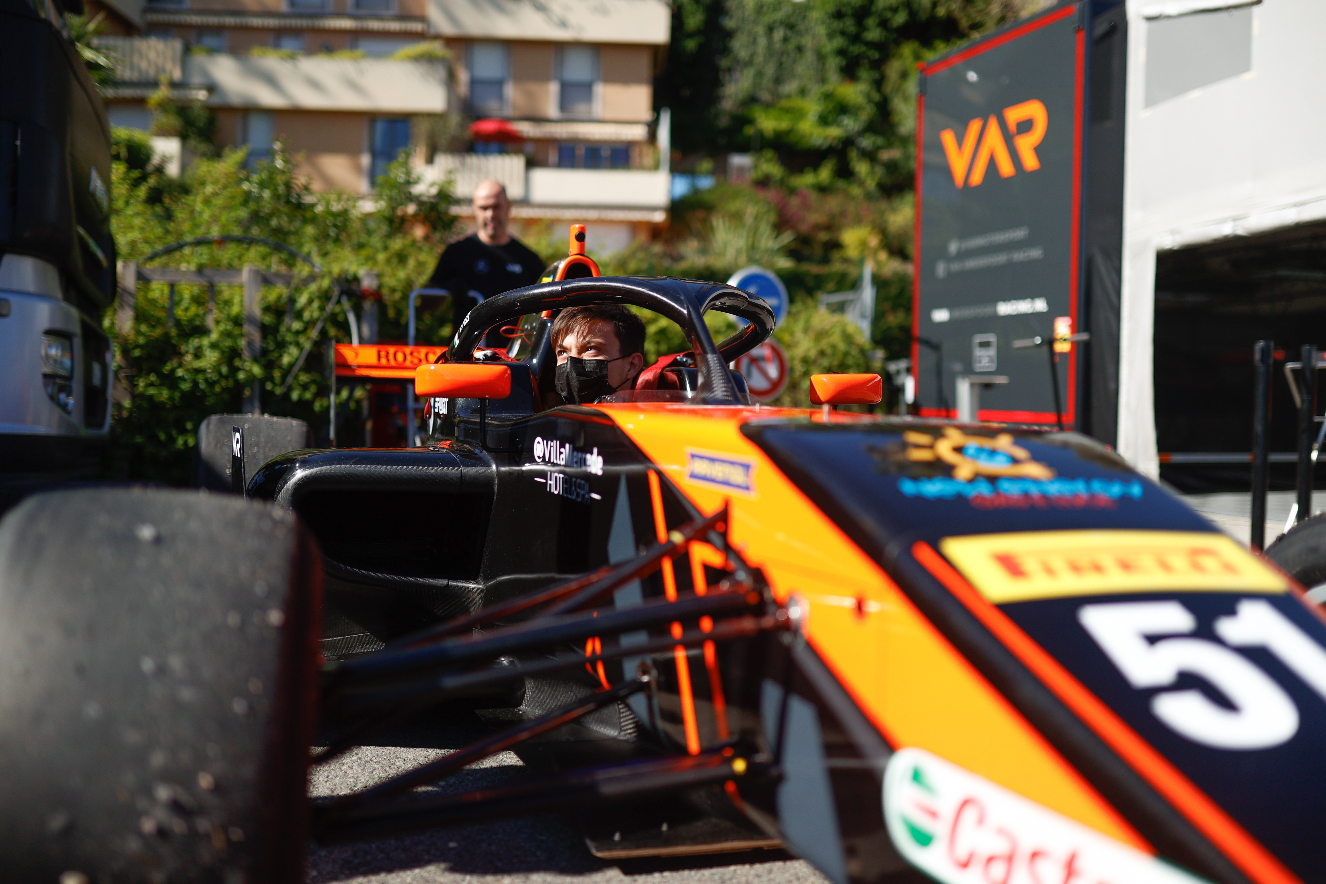 A boy in a mask sits in a black, orange and grey car belonging to Van Amersfoort Racing, whose team trailer is in the background to the right.