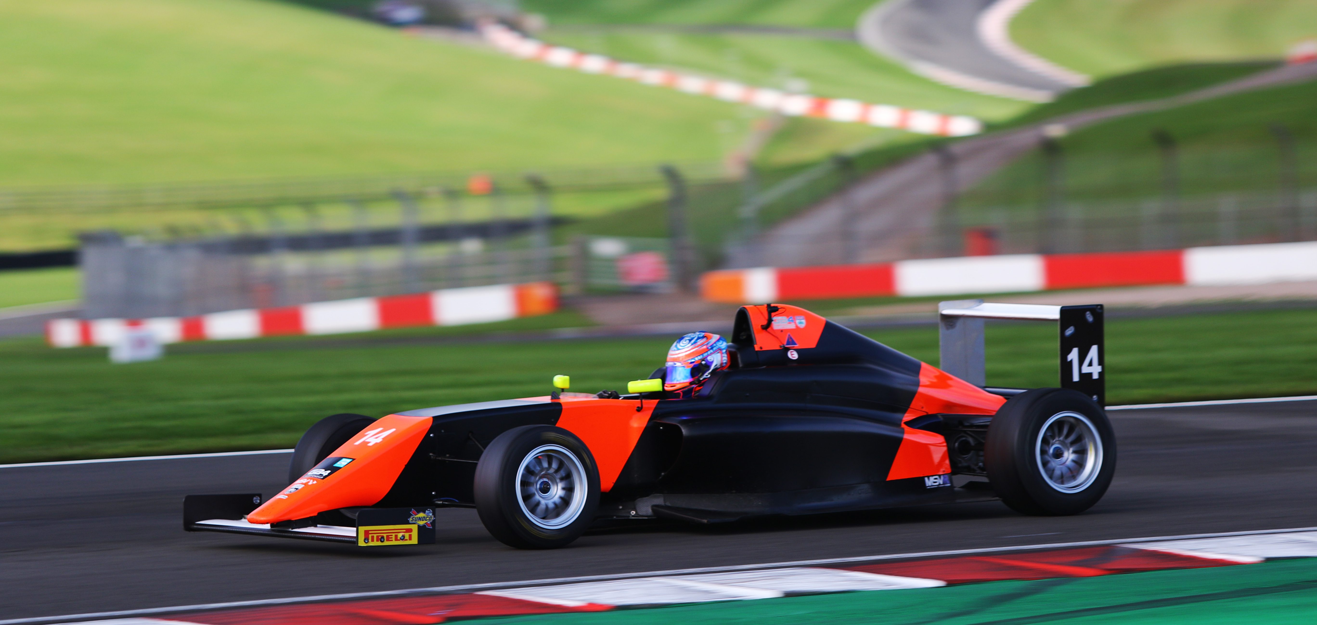 A neon orange and black racecar photographed from the left side on a racetrack with a red and white kerb in the foreground and grass in the background