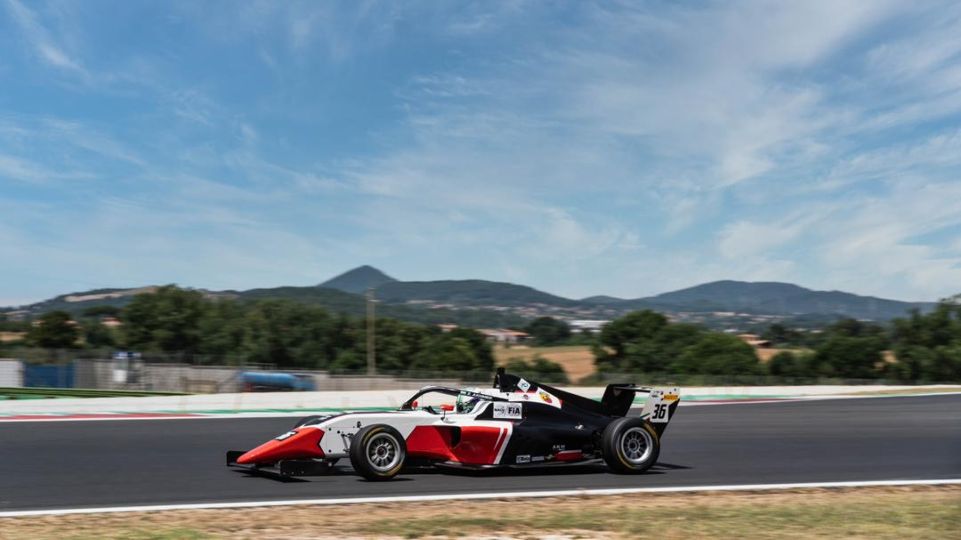 Red and black formula 4 car driving to the left of the camera with mountains in the background.