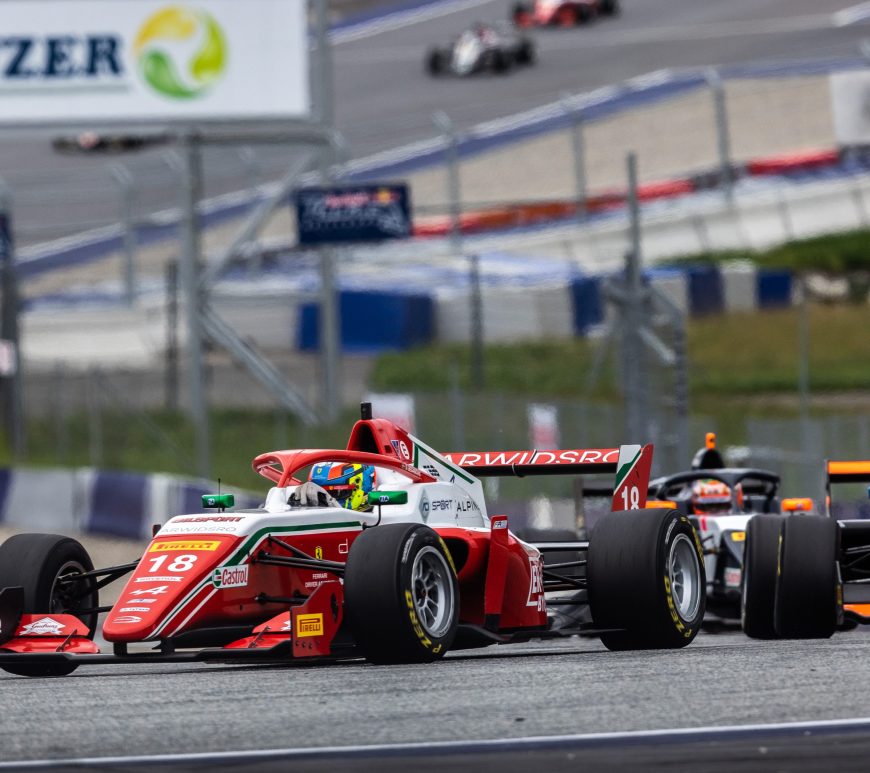 Red and white formula 3 car driving to the left of the camera with many cars behind it.