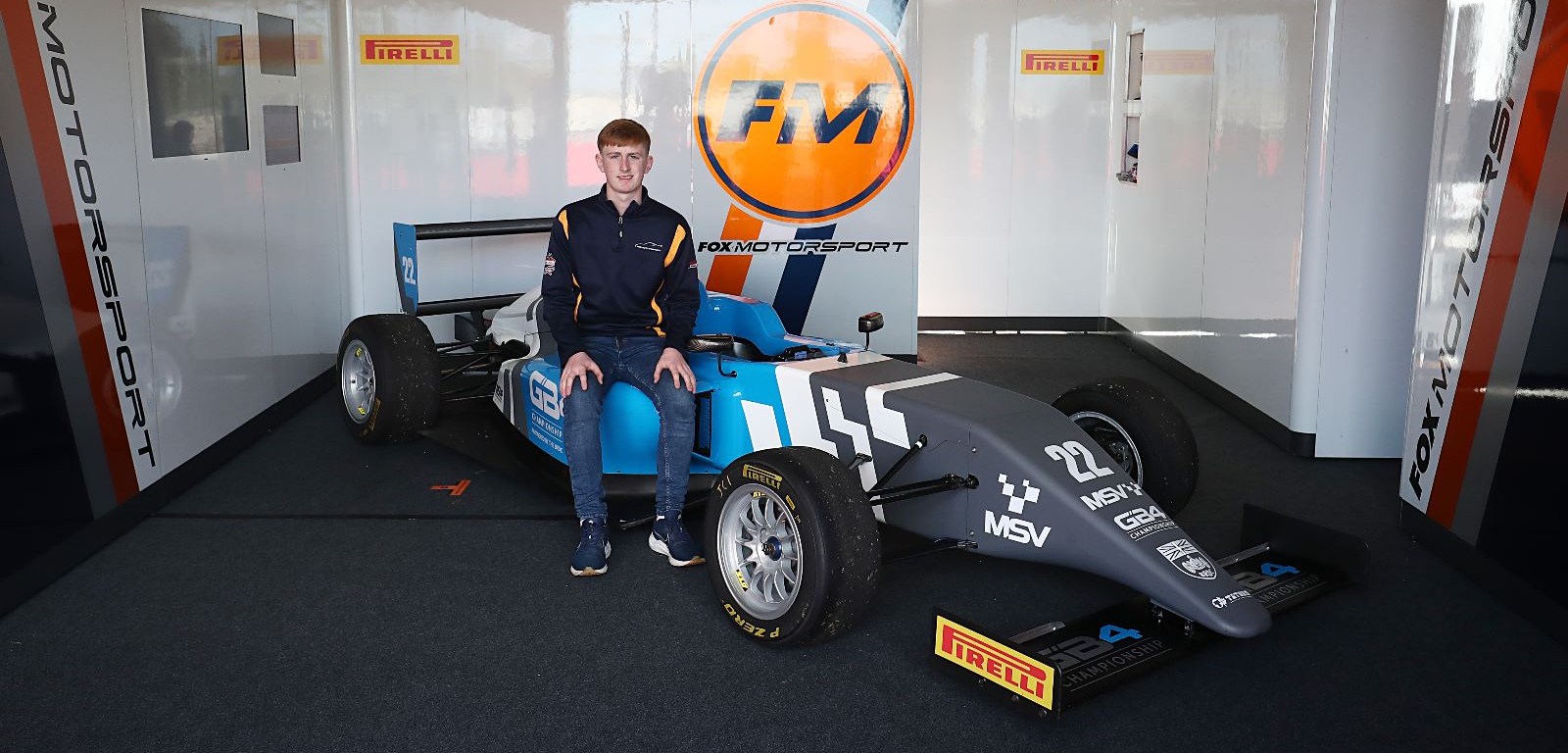 A young man with ginger hair and a blue quarter-zip (Liam McNeilly) sits on a grey, blue and white racecar in a garage with the orange, white and navy blue Fox Motorsport logo on the wall
