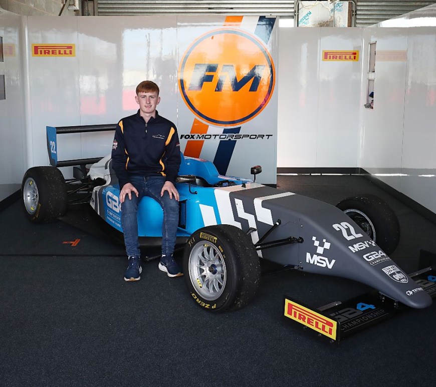 A young man with ginger hair and a blue quarter-zip (Liam McNeilly) sits on a grey, blue and white racecar in a garage with the orange, white and navy blue Fox Motorsport logo on the wall
