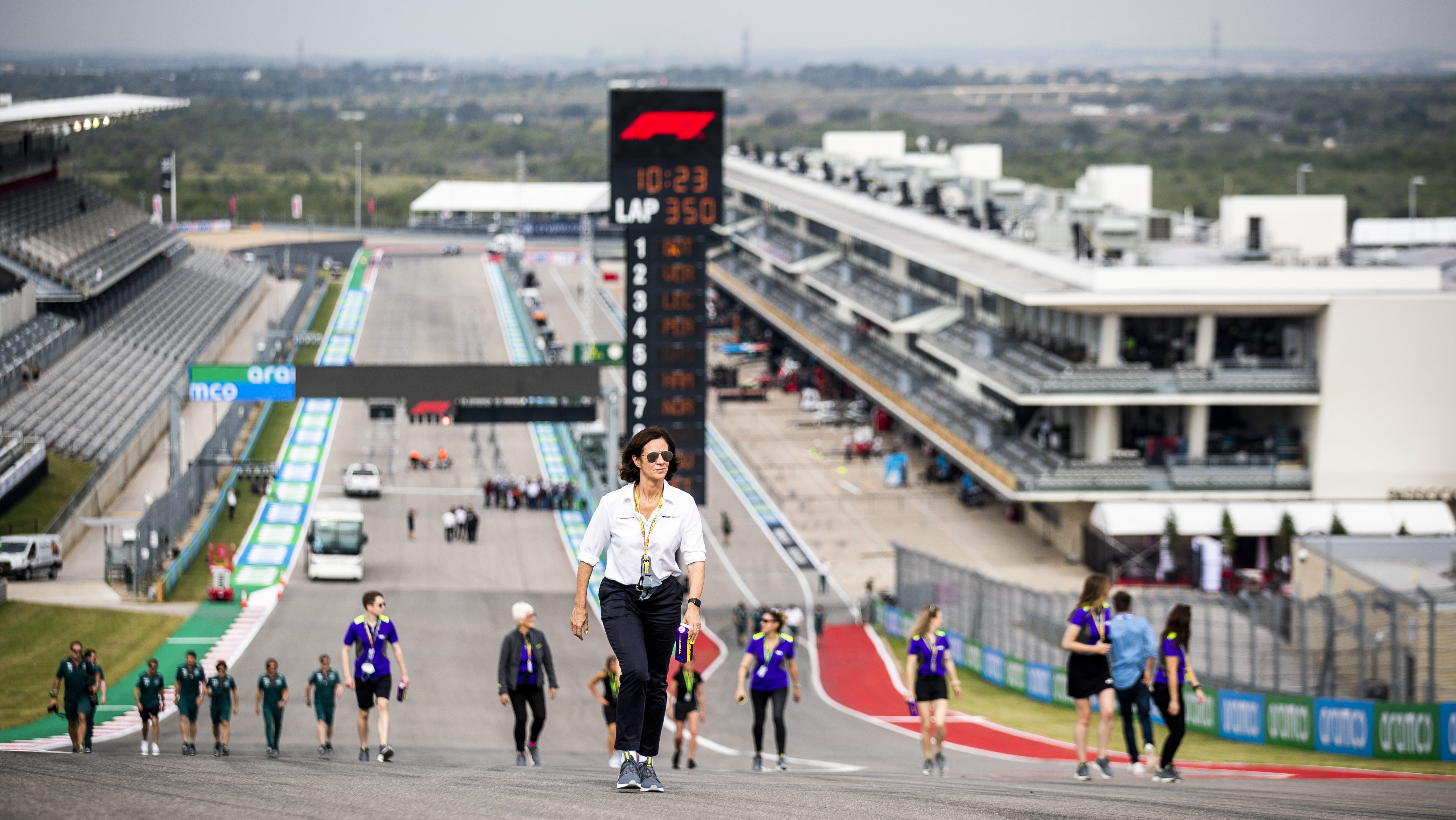 On a cloudy day, a woman (Catherine Bond Muir) in a white shirt and black pants walks up a steep hill on a race track with a timing tower and paddock buildings in the background and several others following her