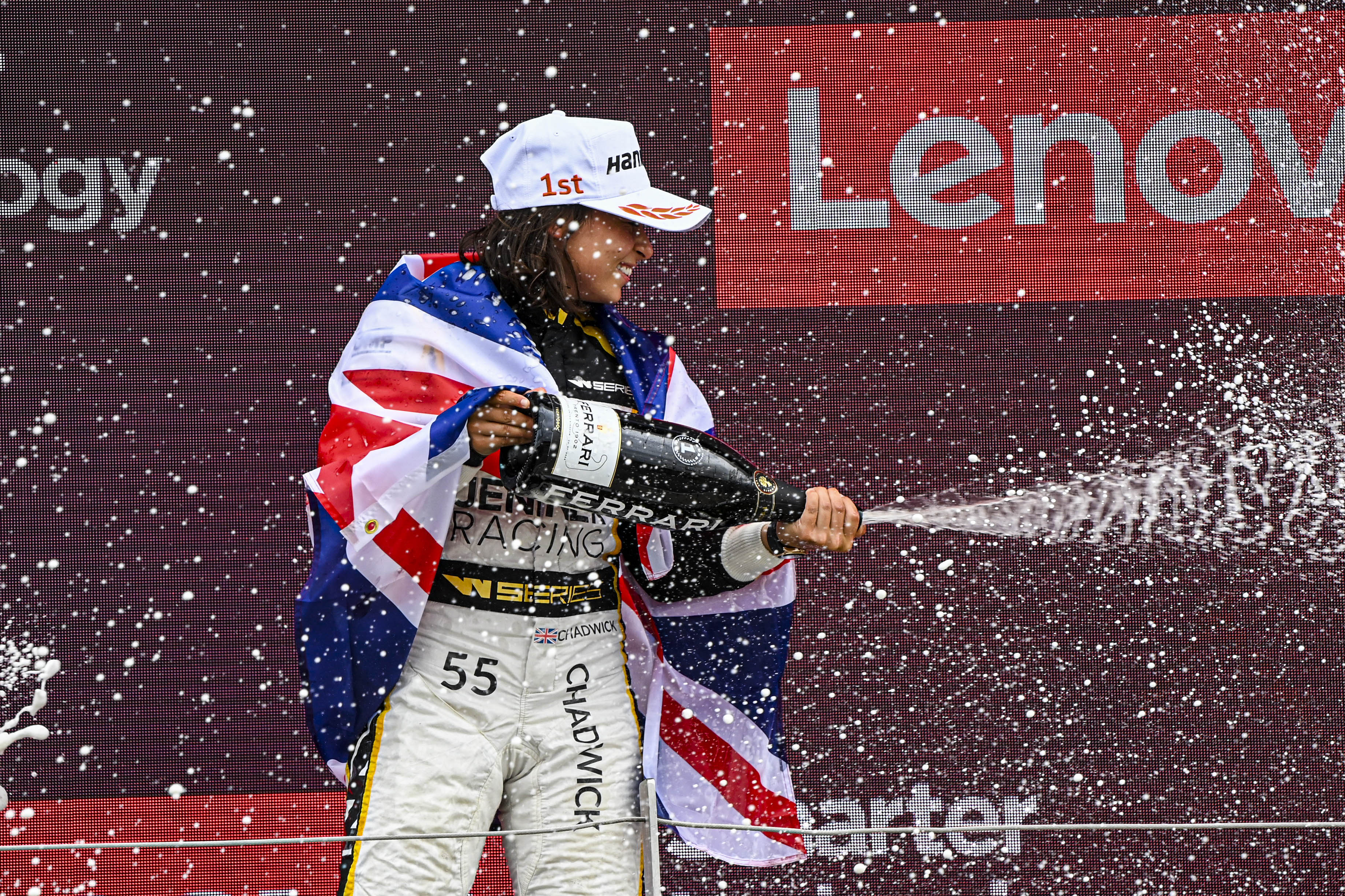 A woman with a white Hankook cap and brown hair sprays sparkling wine on the podium while draped in the Union Jack