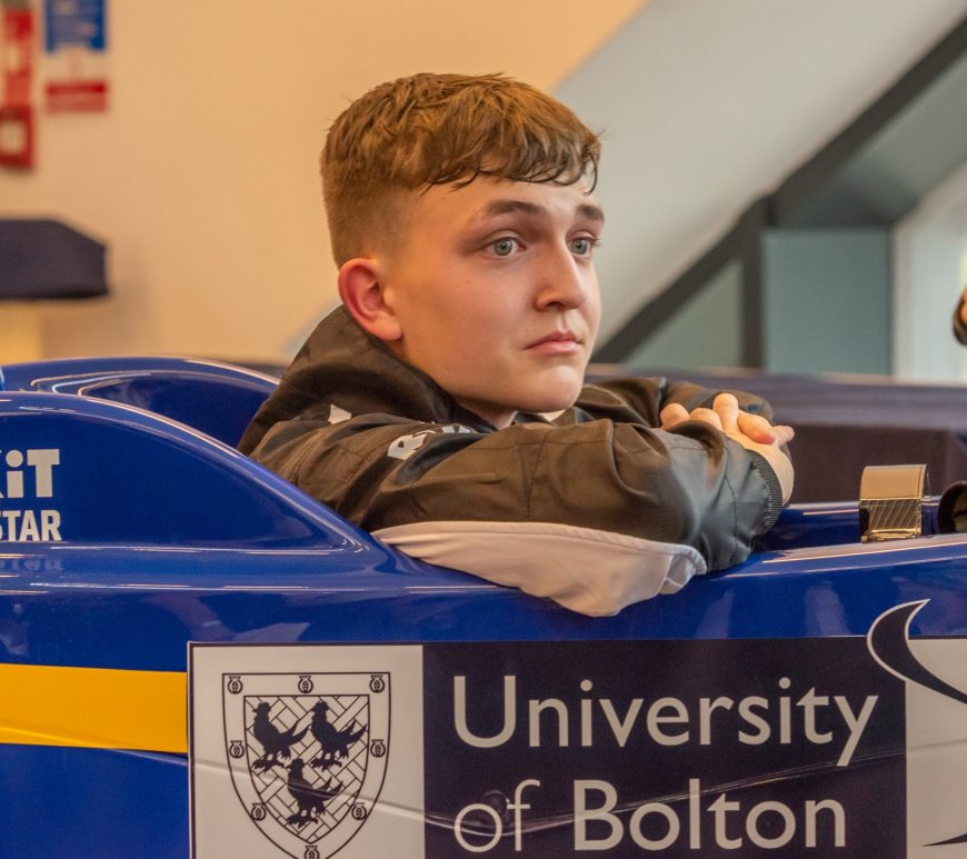 Deagen Fairclough, wearing a race suit, sits in a blue racecar with a yellow stripe in the lower corner and ROKiT Racing Star and University of Bolton branding around the cockpit. His arms are on the sides of the car and he is looking out to his right, slightly angled towards the camera.