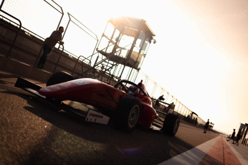 A red Spanish F4 racecar drives past the camera, which is tilted skyward and to the left, in the pit lane of the MotorLand Aragón circuit. The flag tower is in the background along the pit wall, where a few staff members roam and stand by.