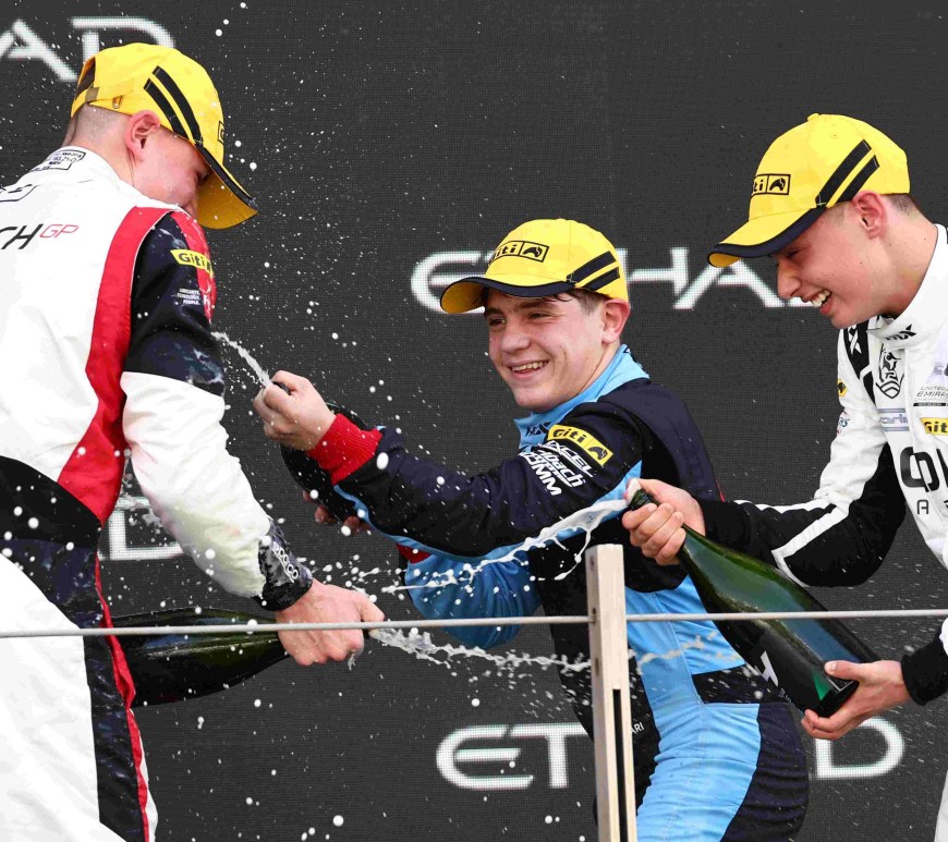Three boys in racing suits and yellow Giti hats spray rose water on one another against a Black podium backdrop with Etihad lettering.