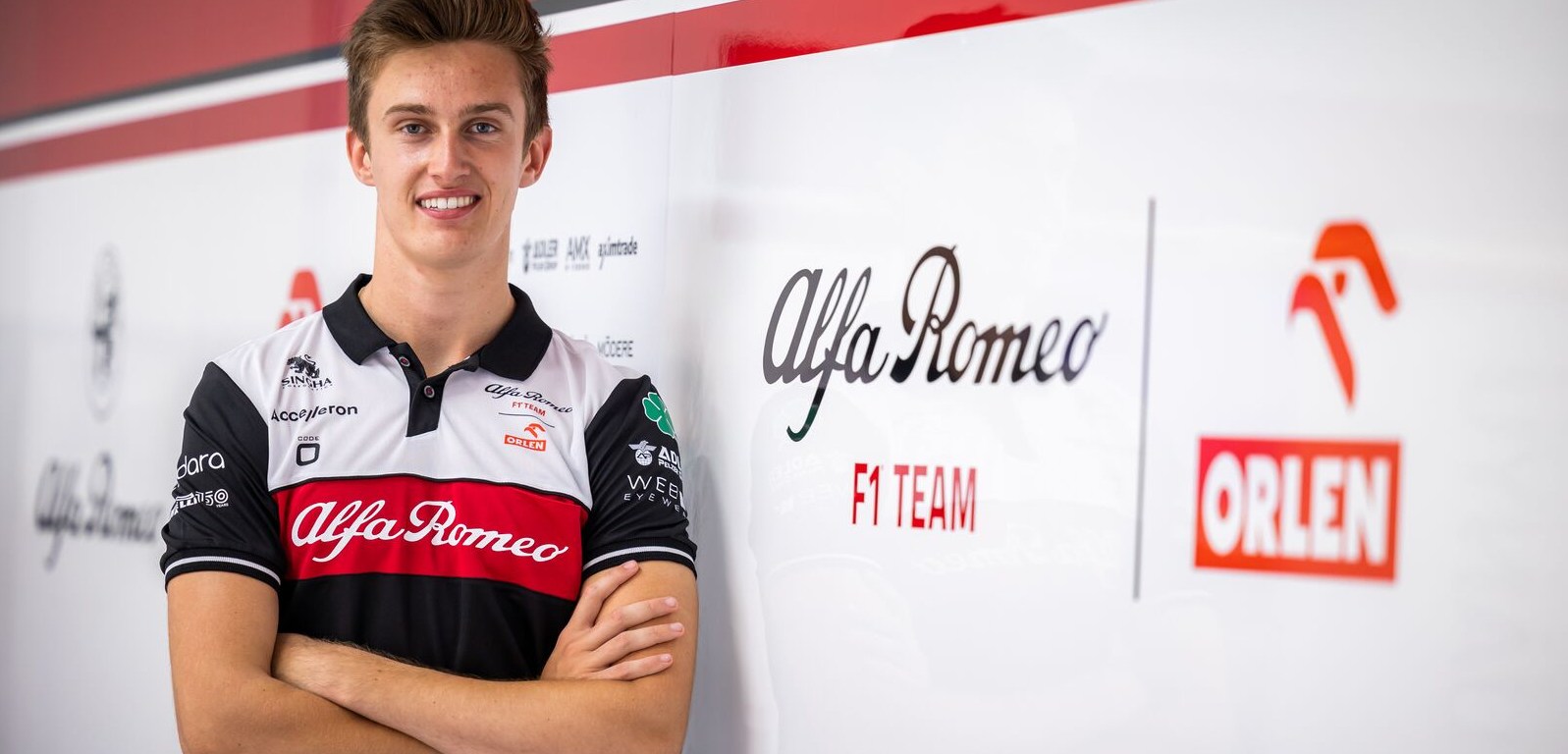 Théo Pourchaire, wearing an Alfa Romeo team shirt and crossing his arms, faces the camera and smiles. An Alfa Romeo F1 Team Orlen logo is on the wall to his right, and Alfa Romeo team sponsor logos blend into the background behind him.