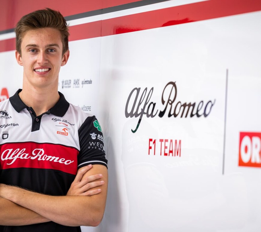 Théo Pourchaire, wearing an Alfa Romeo team shirt and crossing his arms, faces the camera and smiles. An Alfa Romeo F1 Team Orlen logo is on the wall to his right, and Alfa Romeo team sponsor logos blend into the background behind him.