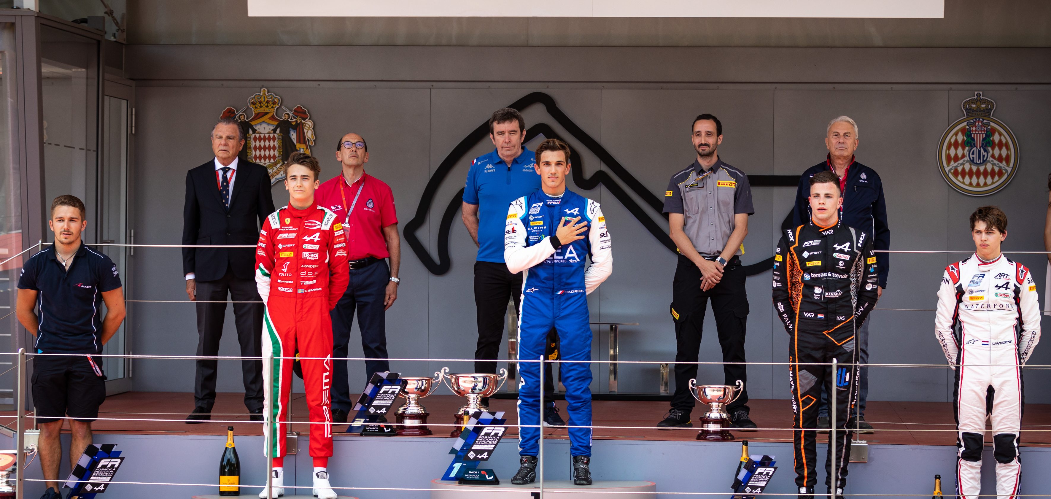 The podium at Monaco. In the center stands Hadrien David in a blue and white racing overall with his right hand over his heart
