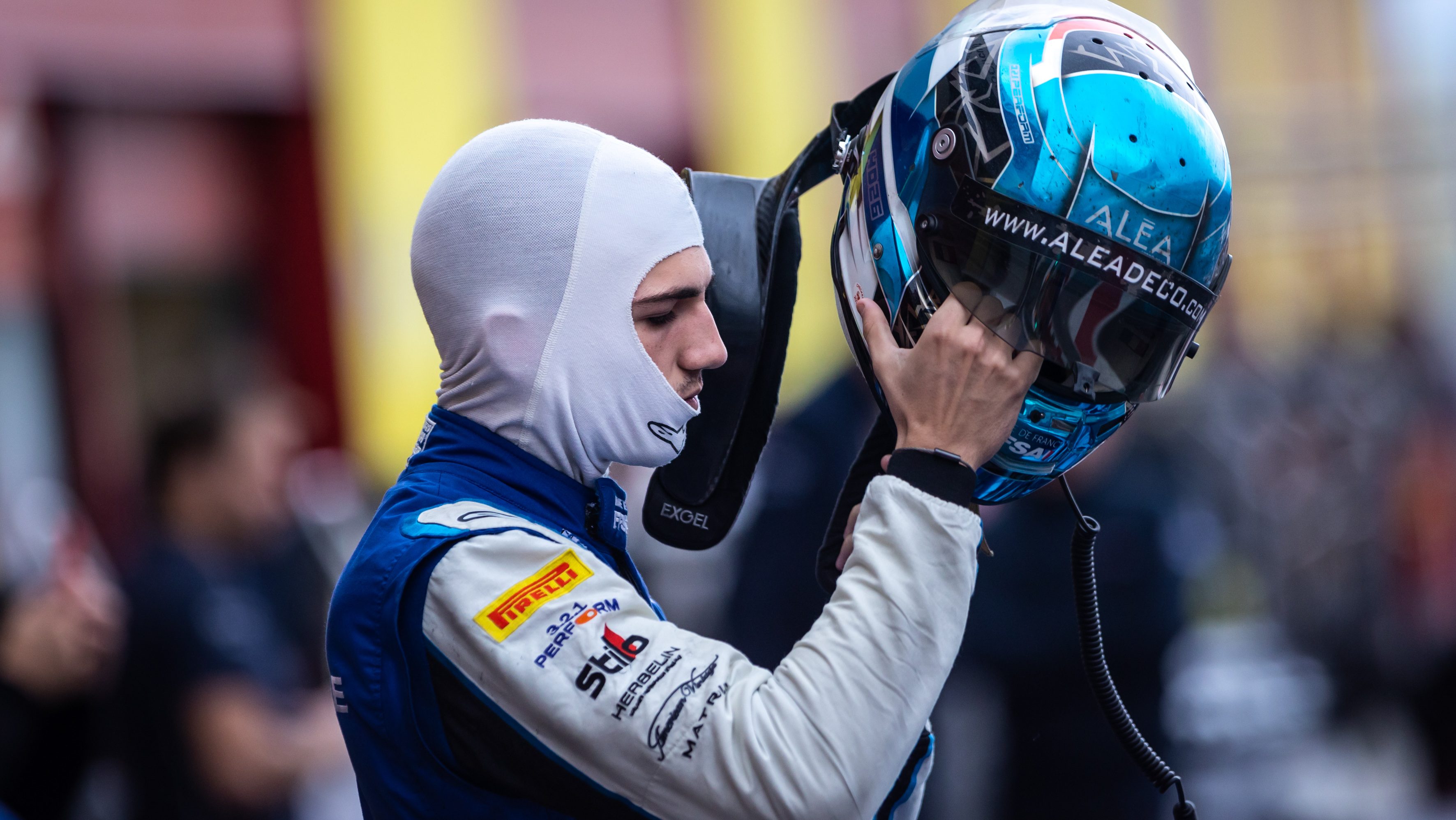 A man in a white balaclava and blue and white racing overalls stands profile to the camera, holding a blue racing helmet aloft in his right hand