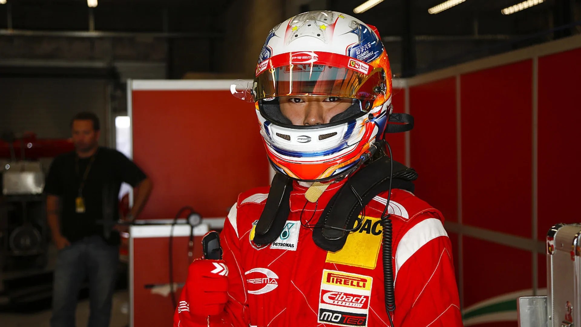 A boy in a white and red helmet adorned with Prema and Ferrari Driver Academy logos and a red racing suit gives a thumbs-up gesture to the camera