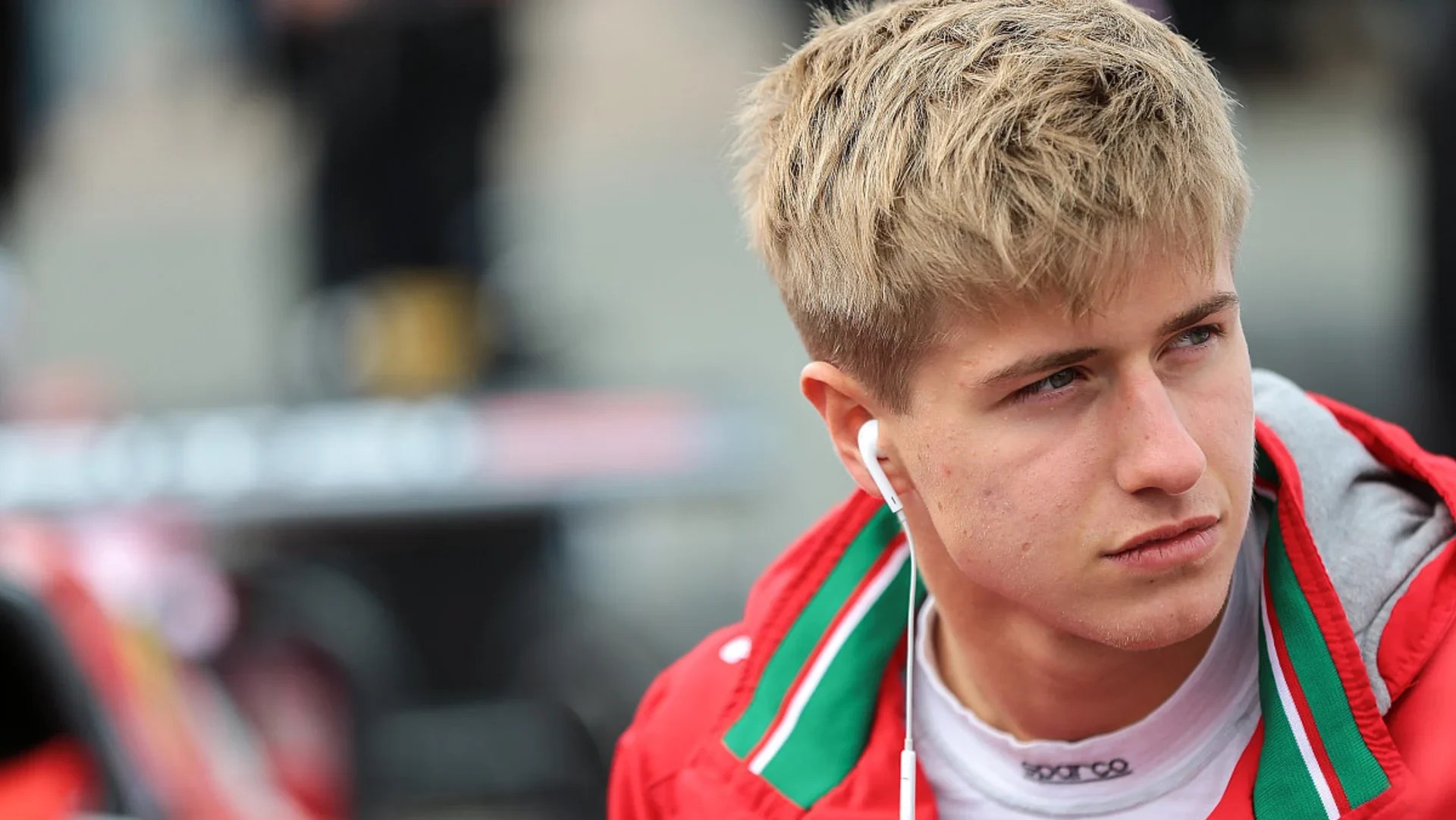 A boy with blond hair in a red jacket over a Sparco race suit looks pensively to the left with his right Apple EarPod in his ear