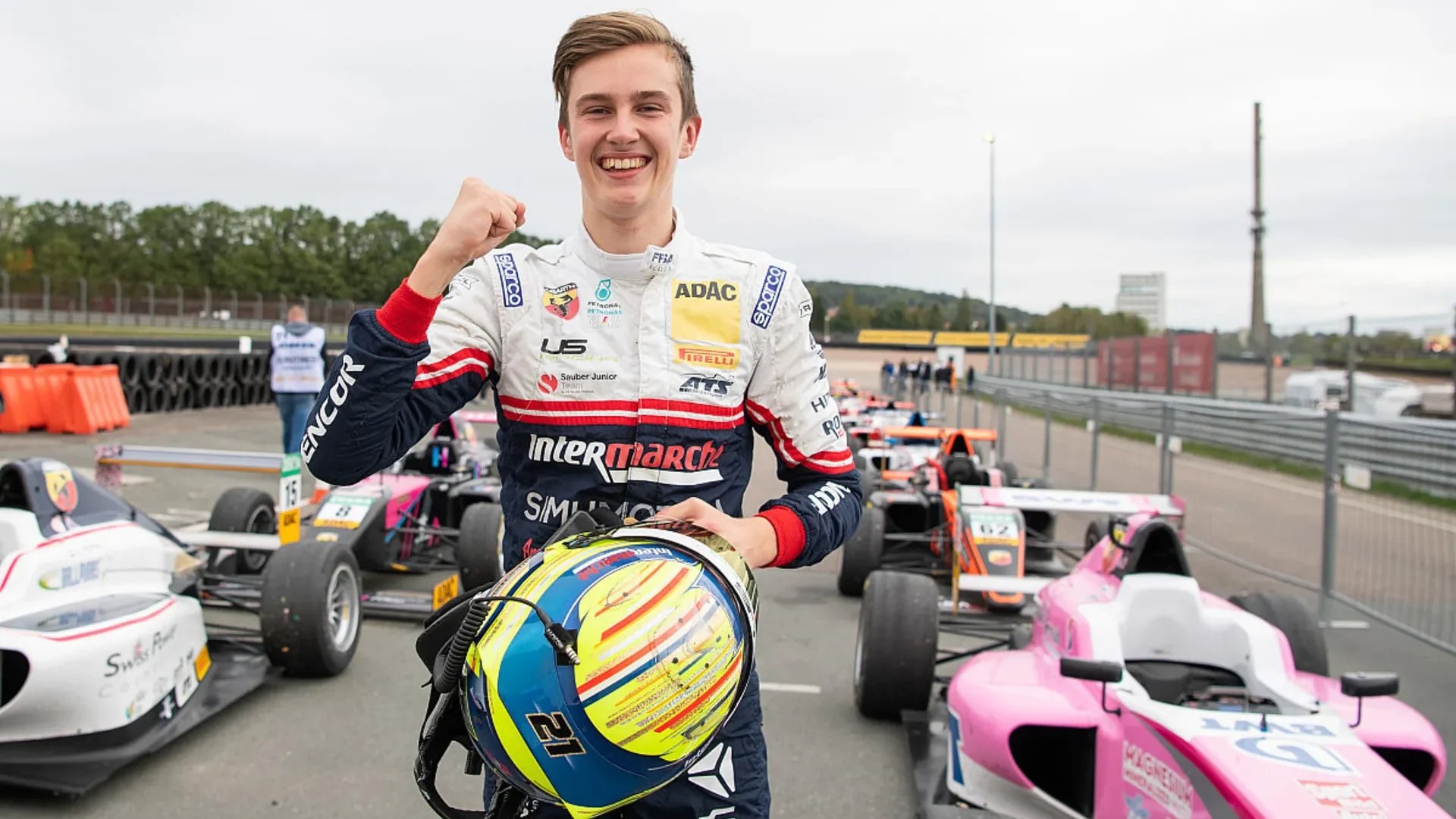 A boy in a white and blue racing suit holding a yellow and blue helmet smiles at the camera and holds up his right fist. Pink, orange, white and black cars line up behind him two by two.