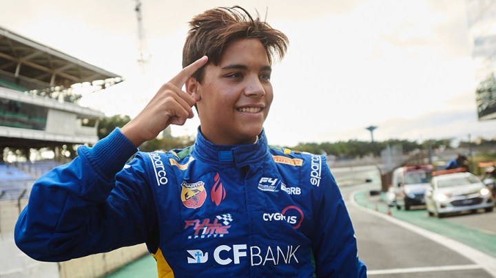 A driver in a blue suit smiles while looking to the left and raises his right index finger