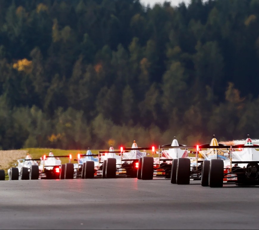 Viewed from the rear, eight racecars are lined up nose to tail on asphalt as green trees cascade behind them in the distance