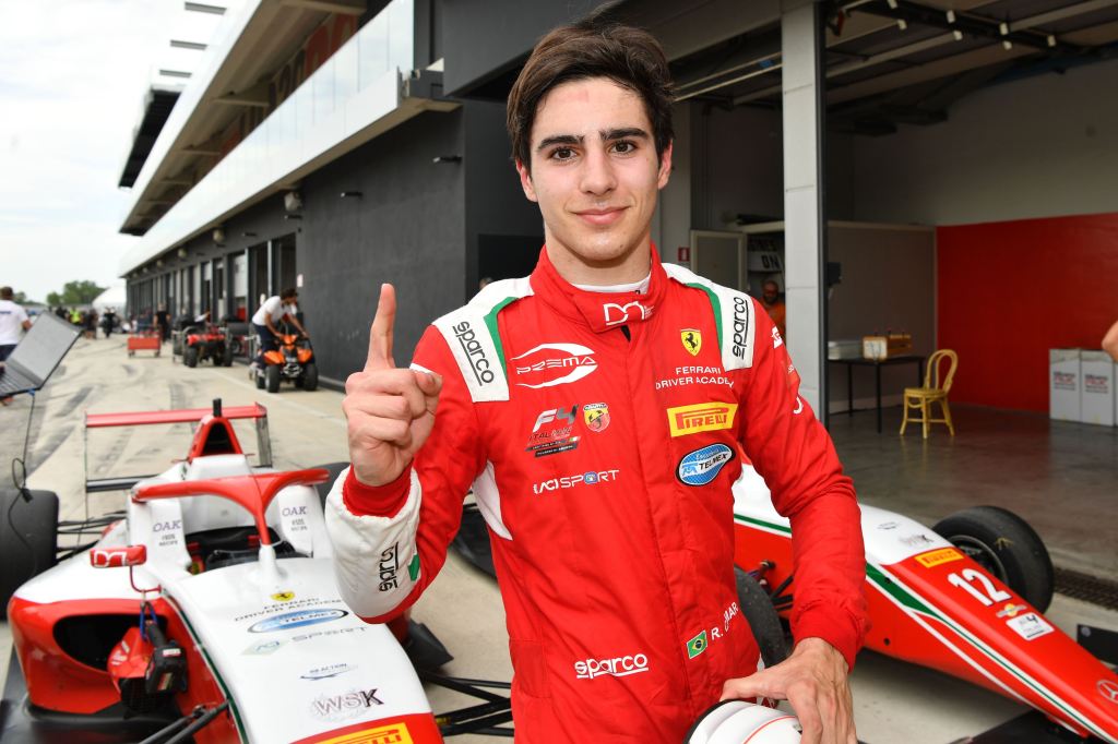 A driver in a red racing suit puts his right index finger up