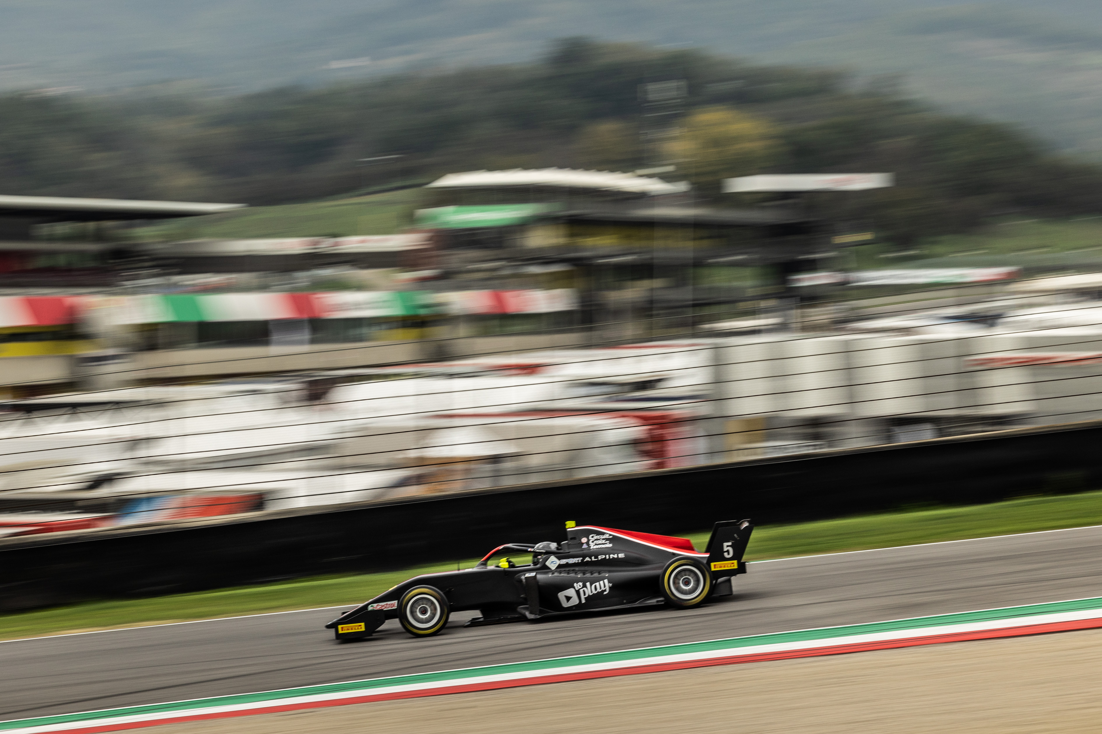A black car with red accents driving past the Italian tricolore and gravel bed nearest the camera with buildings and trucks blurred in the background
