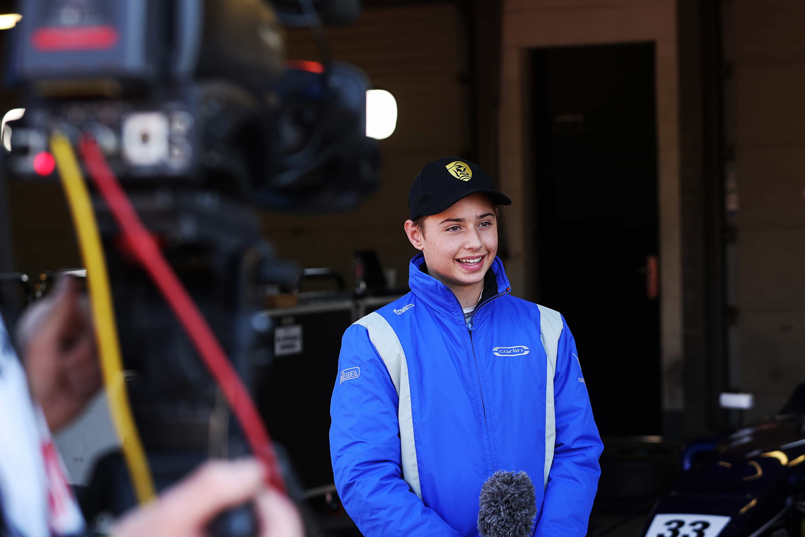 A driver in a blue jacket with a black cap smiles and holds a microphone
