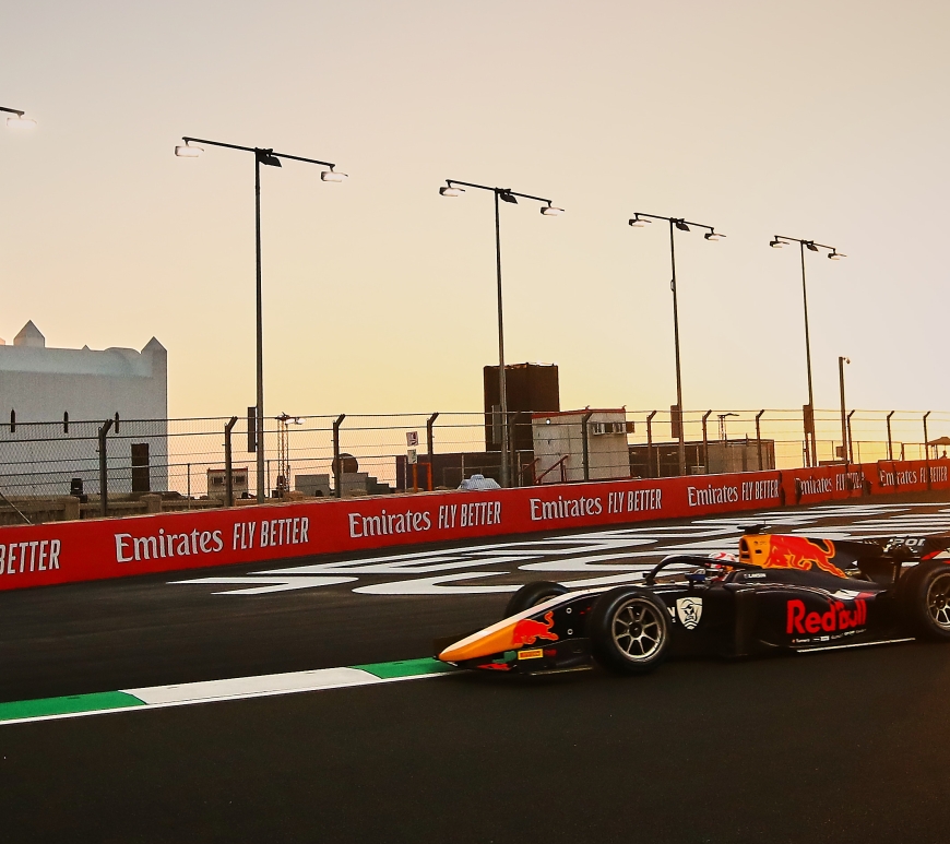 The blue and white Red Bull–liveried F2 car of Liam Lawson negotiates a slight right turn on the Jeddah Corniche Circuit, with the glare of the setting sun illuminating the sky and a mosque visible behind the "Emirates Fly Better" branding on the outside wall.
