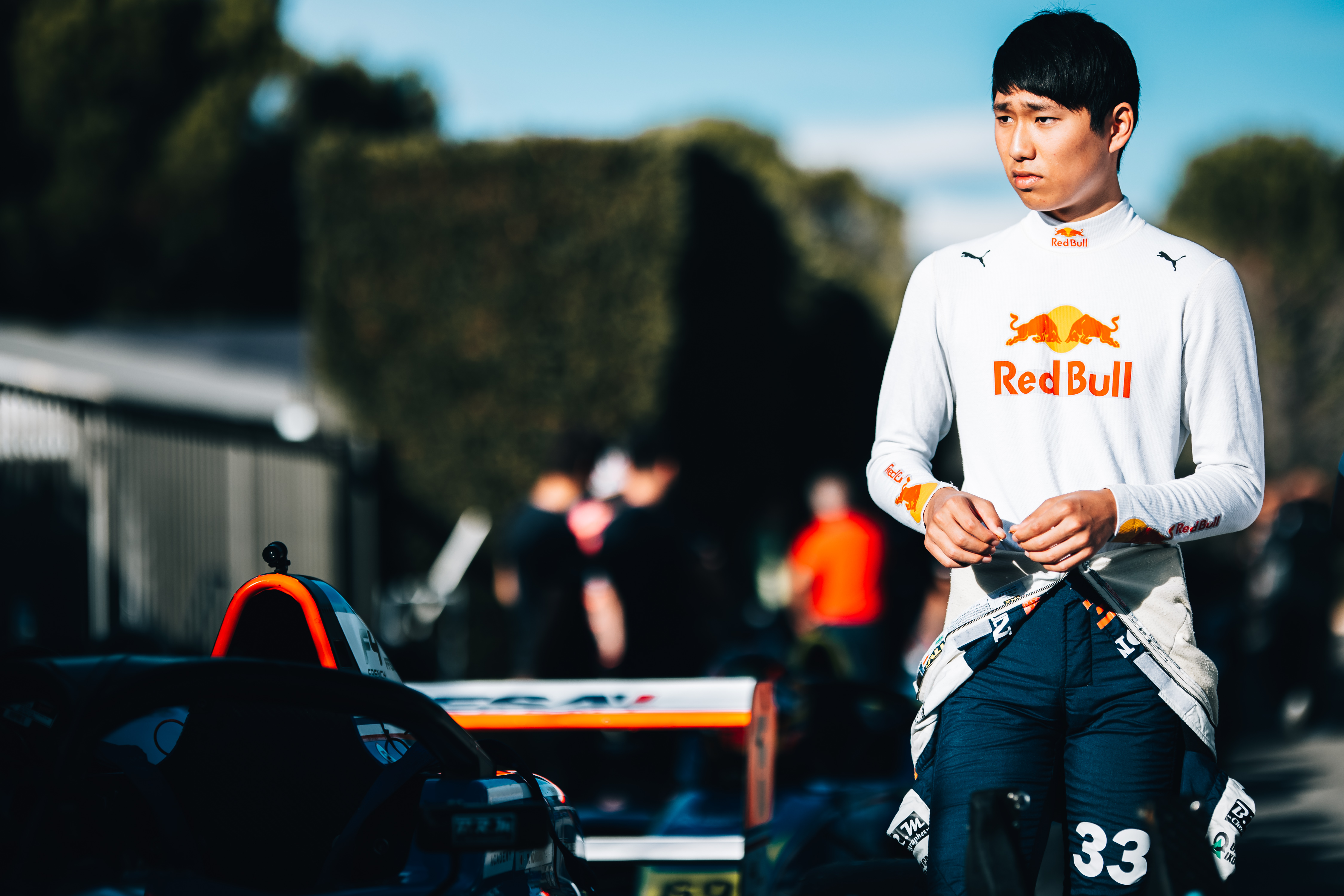A driver in a white race suit with Red Bull branding and overalls at his waist looks to the right