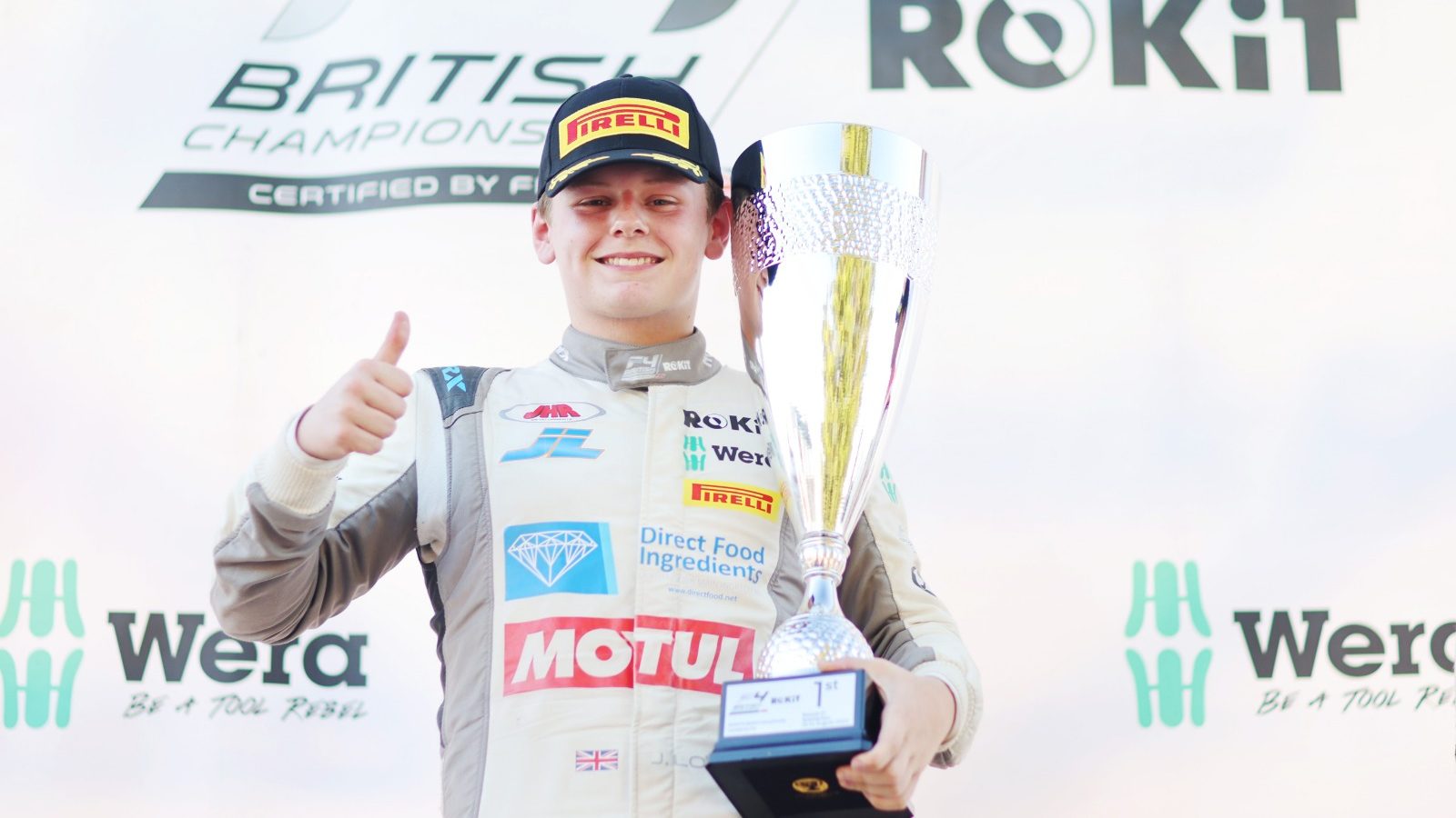 Joseph Loake gives a thumbs-up to the camera and, in his left hand, holds a first-place trophy that rests against his face. ROKiT and Wera logos are displayed in the background.