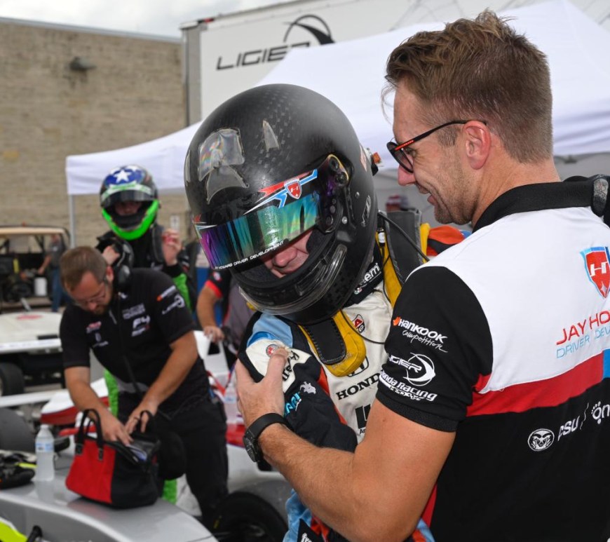 A driver in a black crash helmet hugs a man in a Jay Howard Driver Development polo shirt as several other racing staff look on in the background at parc fermé